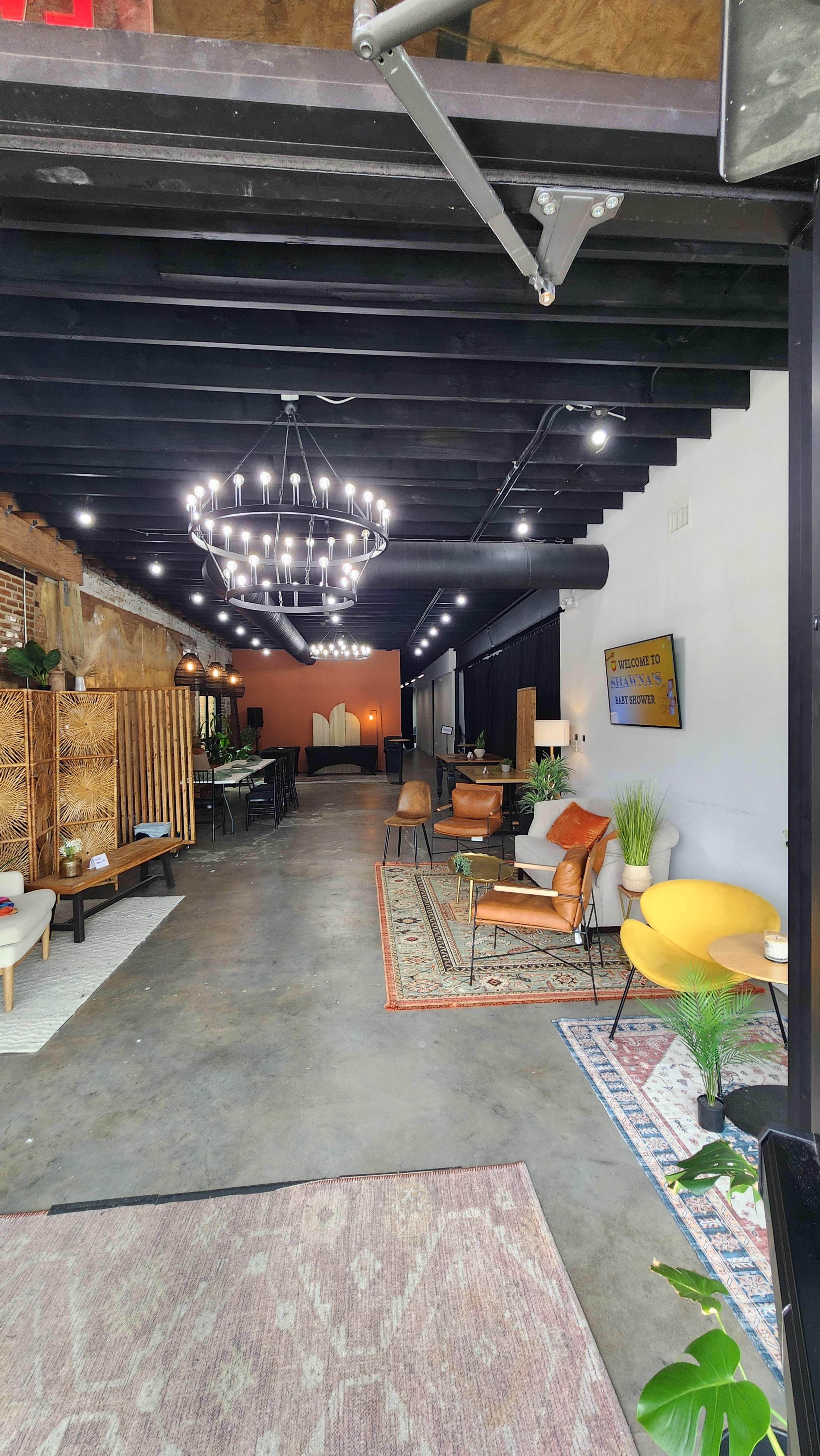 The image shows an interior space with a modern design featuring a chandelier, various seating arrangements, and decorative rugs along with potted plants.