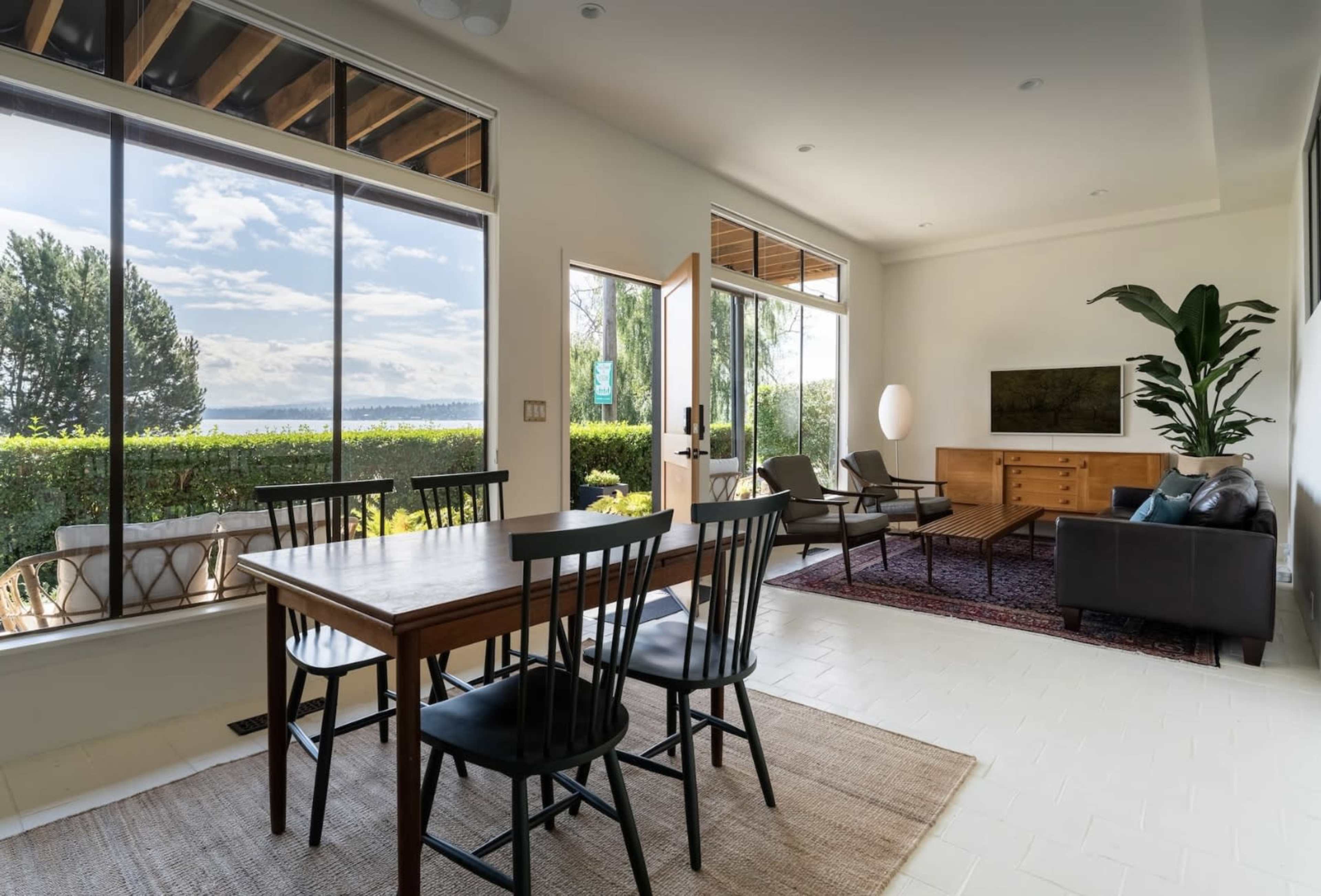 A bright living space features a dining table with black chairs, a seating area with a sofa and chairs, and large windows overlooking a green landscape and water.