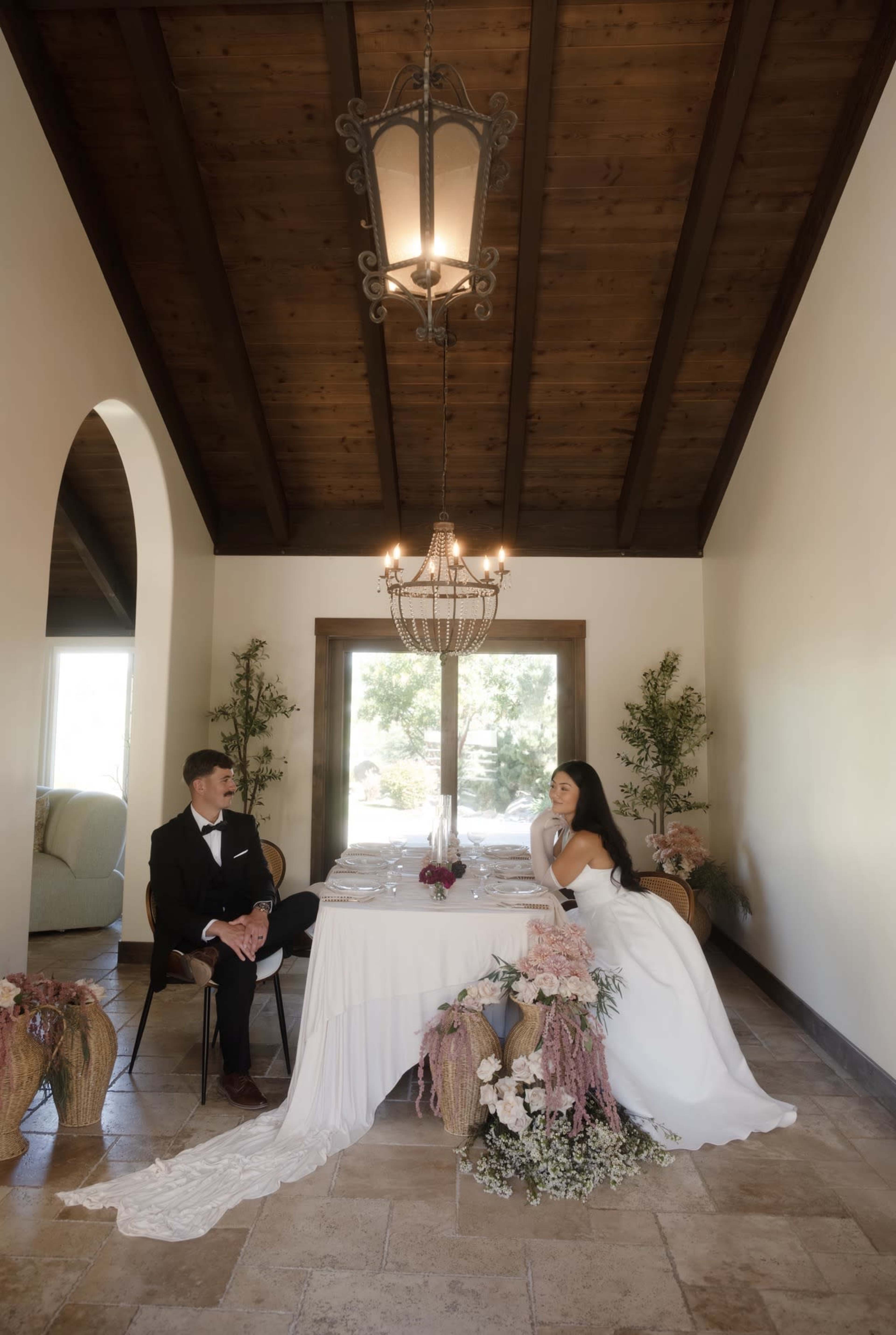 A couple is seated at an elegantly set dining table adorned with floral arrangements in a well-lit room with a wooden ceiling.
