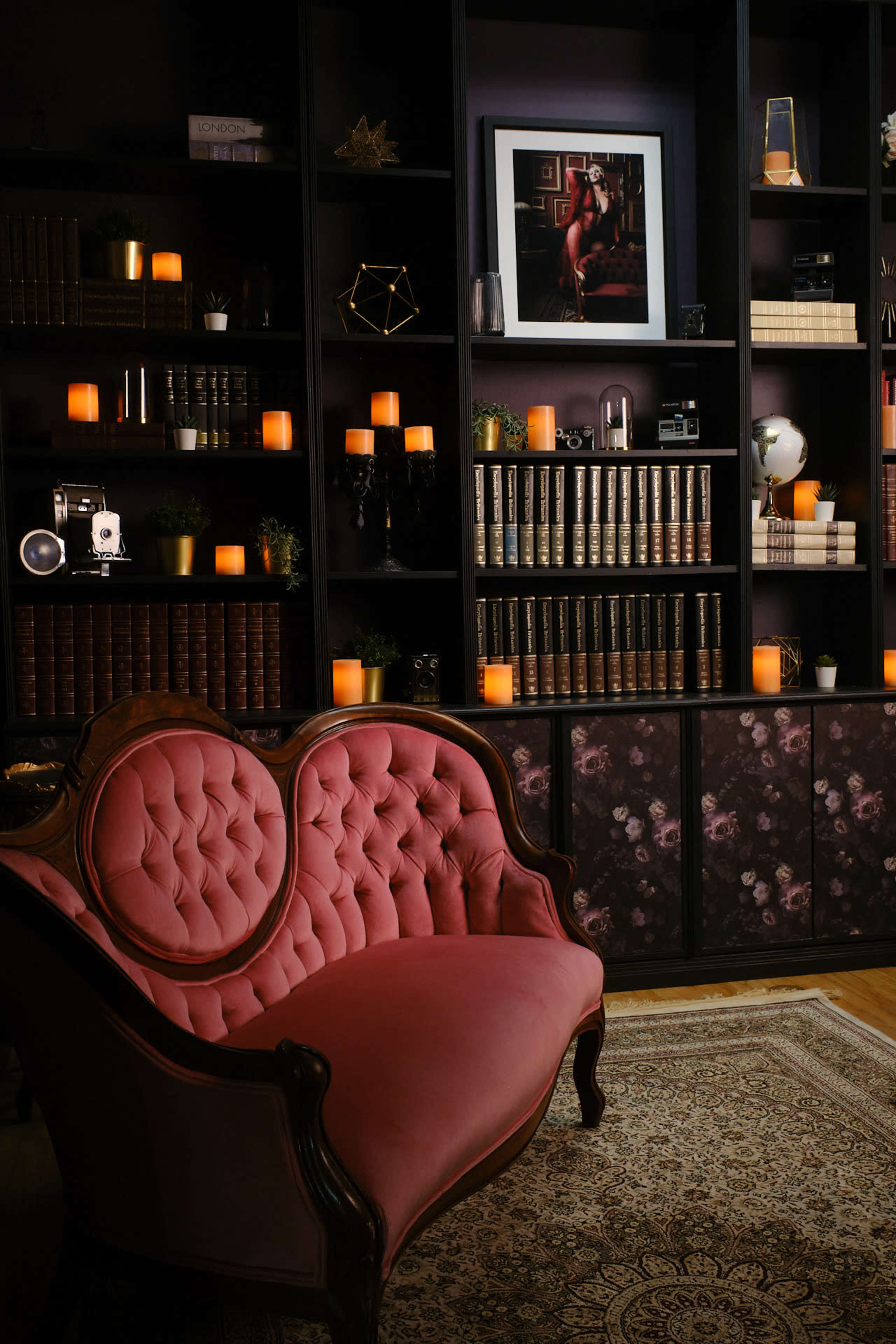 A vintage pink loveseat in front of a dark bookshelf filled with books and decorative items, illuminated by warm candlelight.