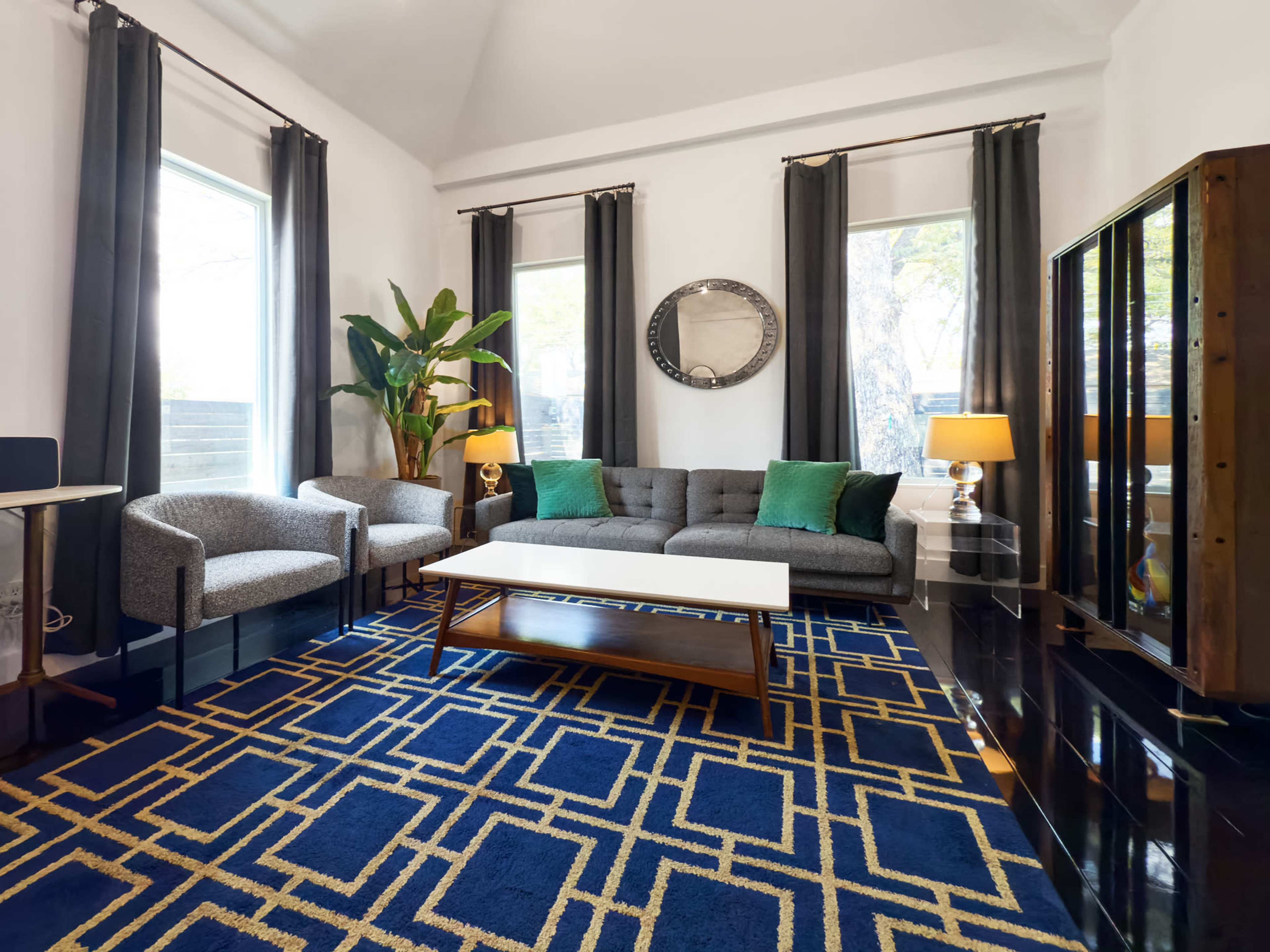 The living room features a gray sofa with green cushions, two patterned chairs, a coffee table, a large blue area rug, and large windows with curtains.