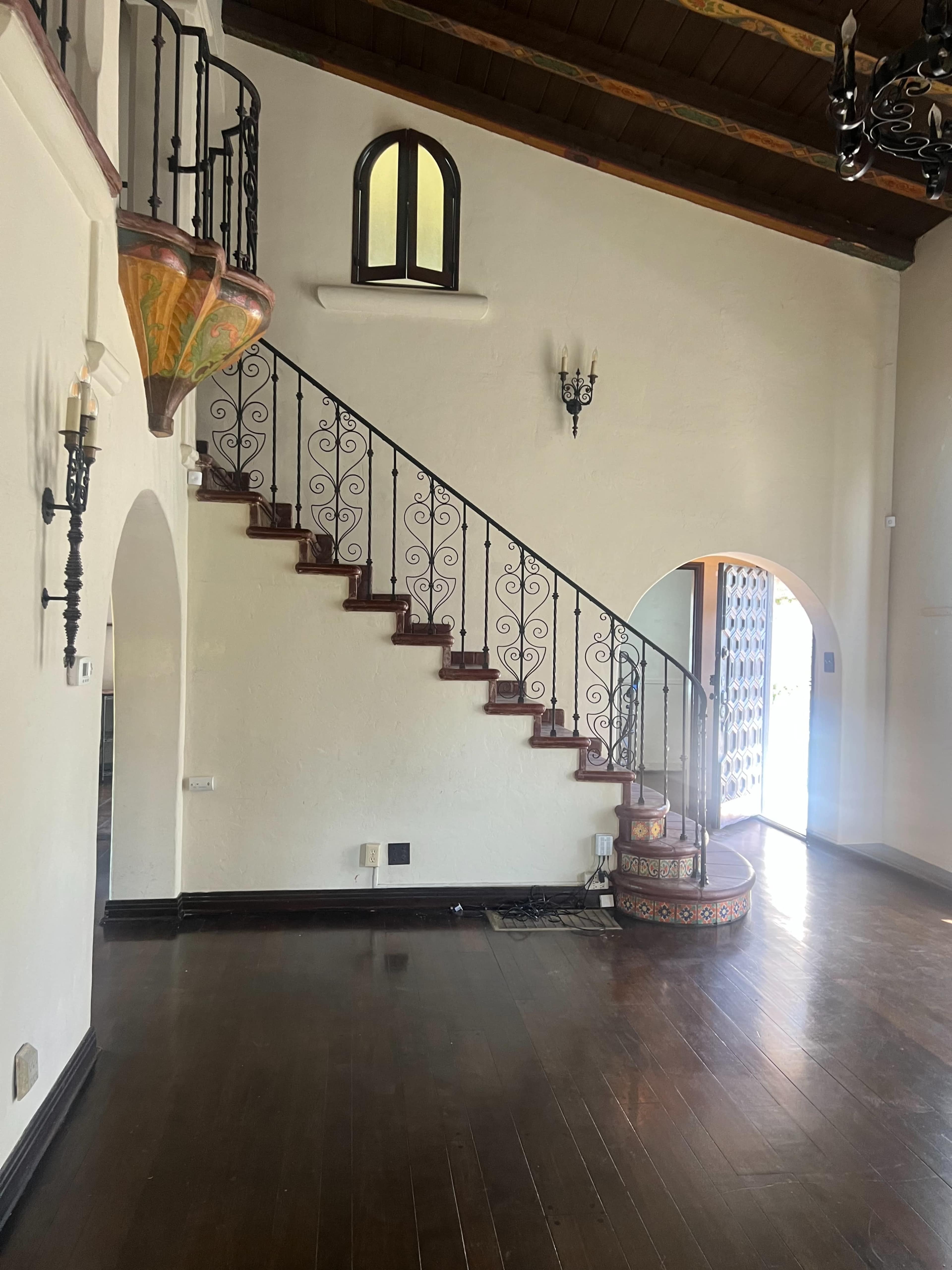 The image shows a well-lit interior space featuring a winding staircase with ornate iron railings and an arched doorway.