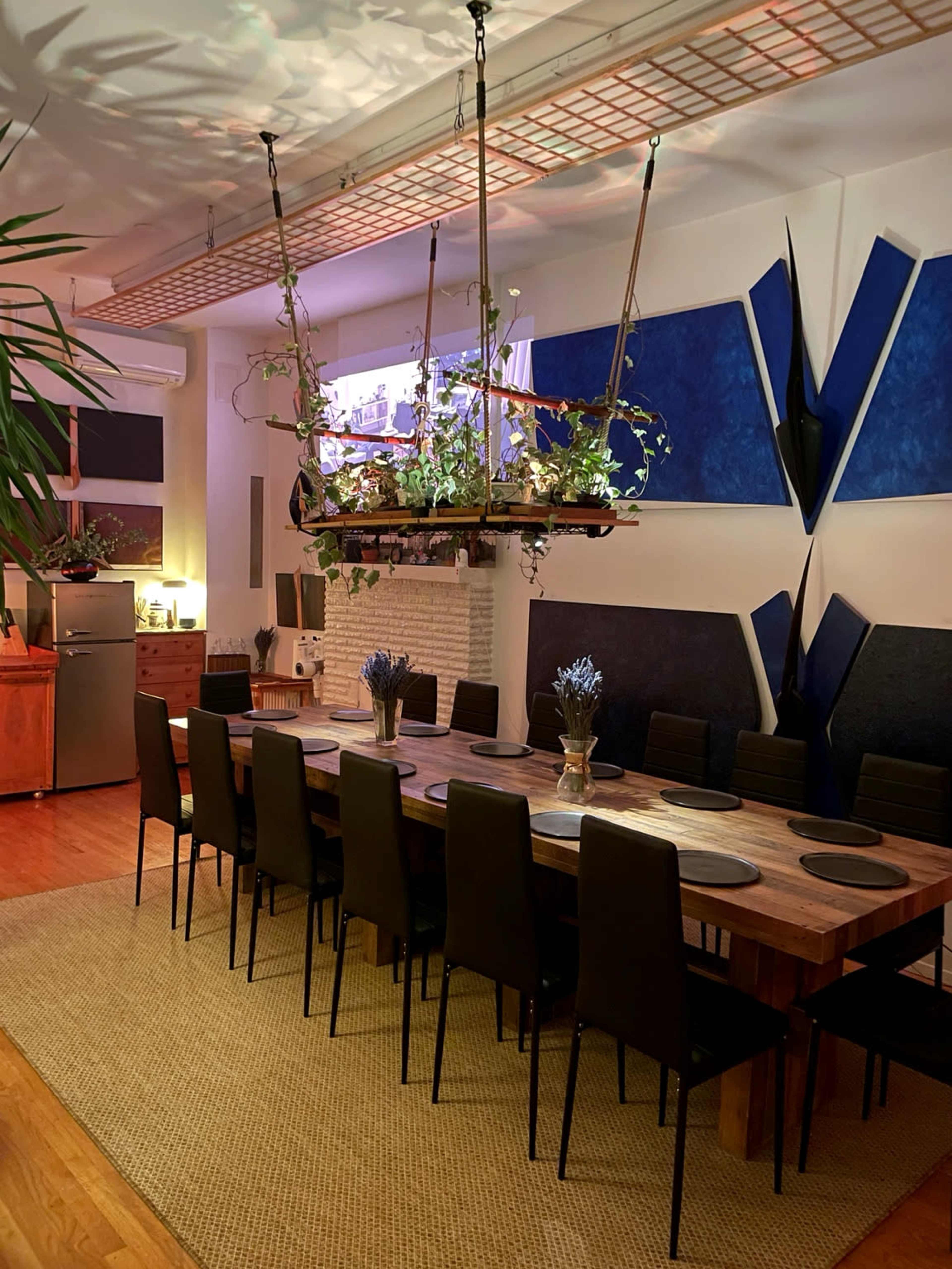 A long dining table is set in a modern room, featuring hanging planters and artistic blue wall decor.