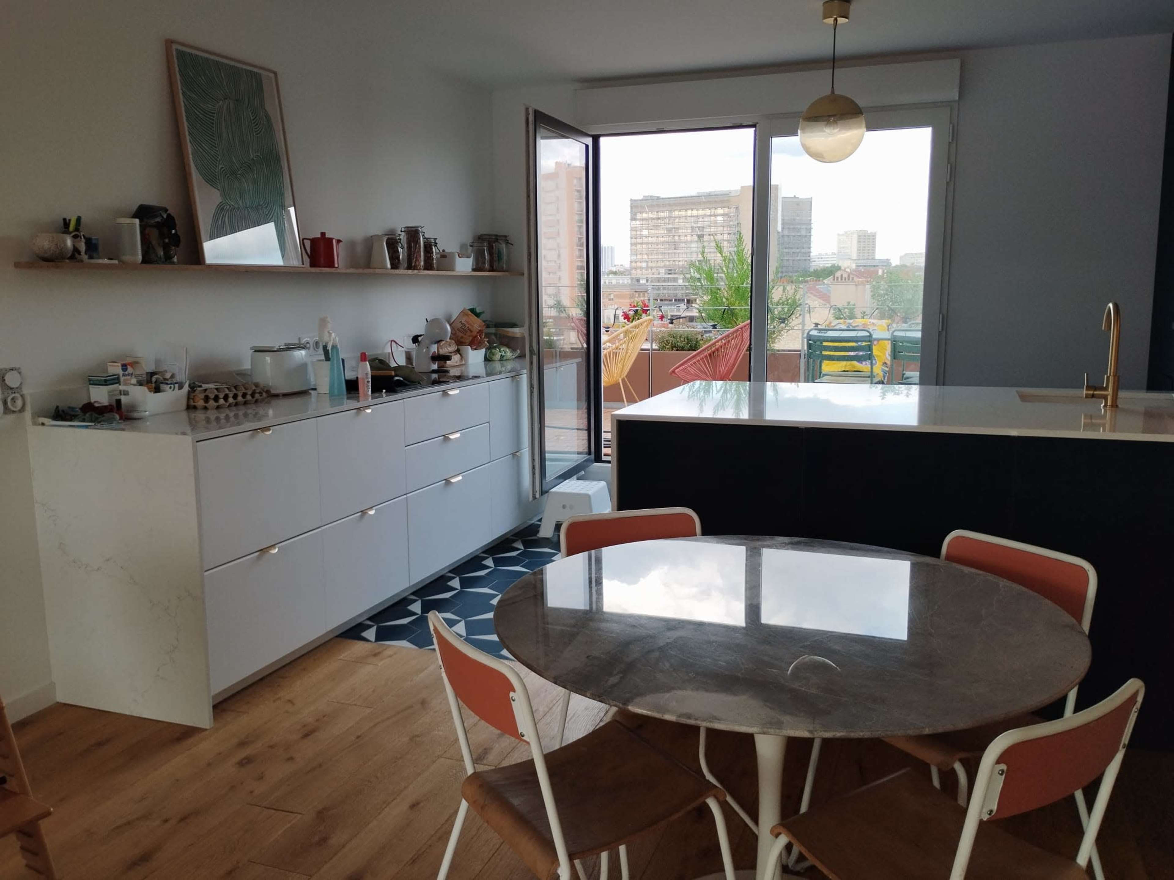 A modern kitchen features a round table with white chairs, a marble-topped counter, and a large window overlooking a cityscape.