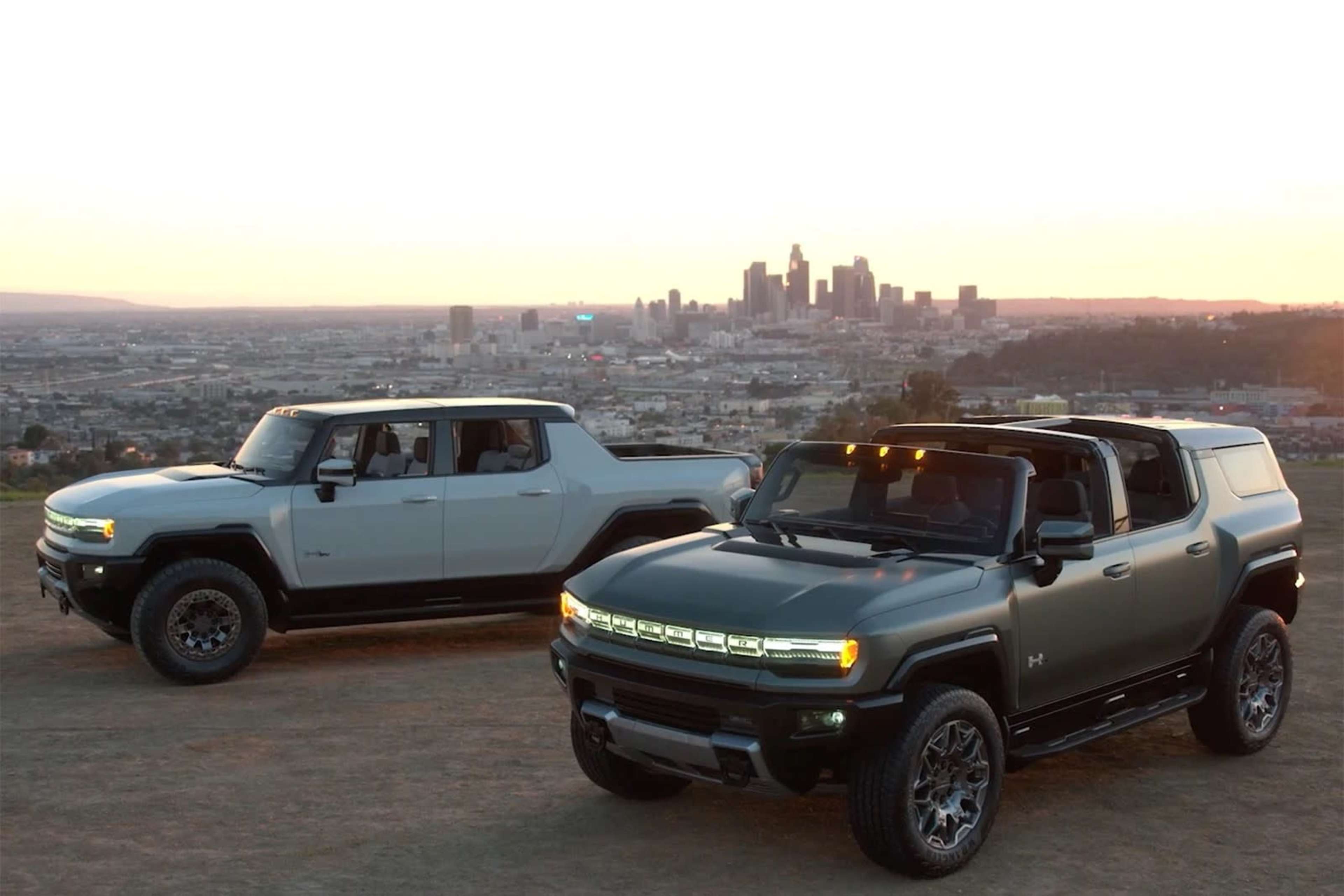 Two Hummer electric trucks are parked on a hillside with a city skyline in the background during sunset.