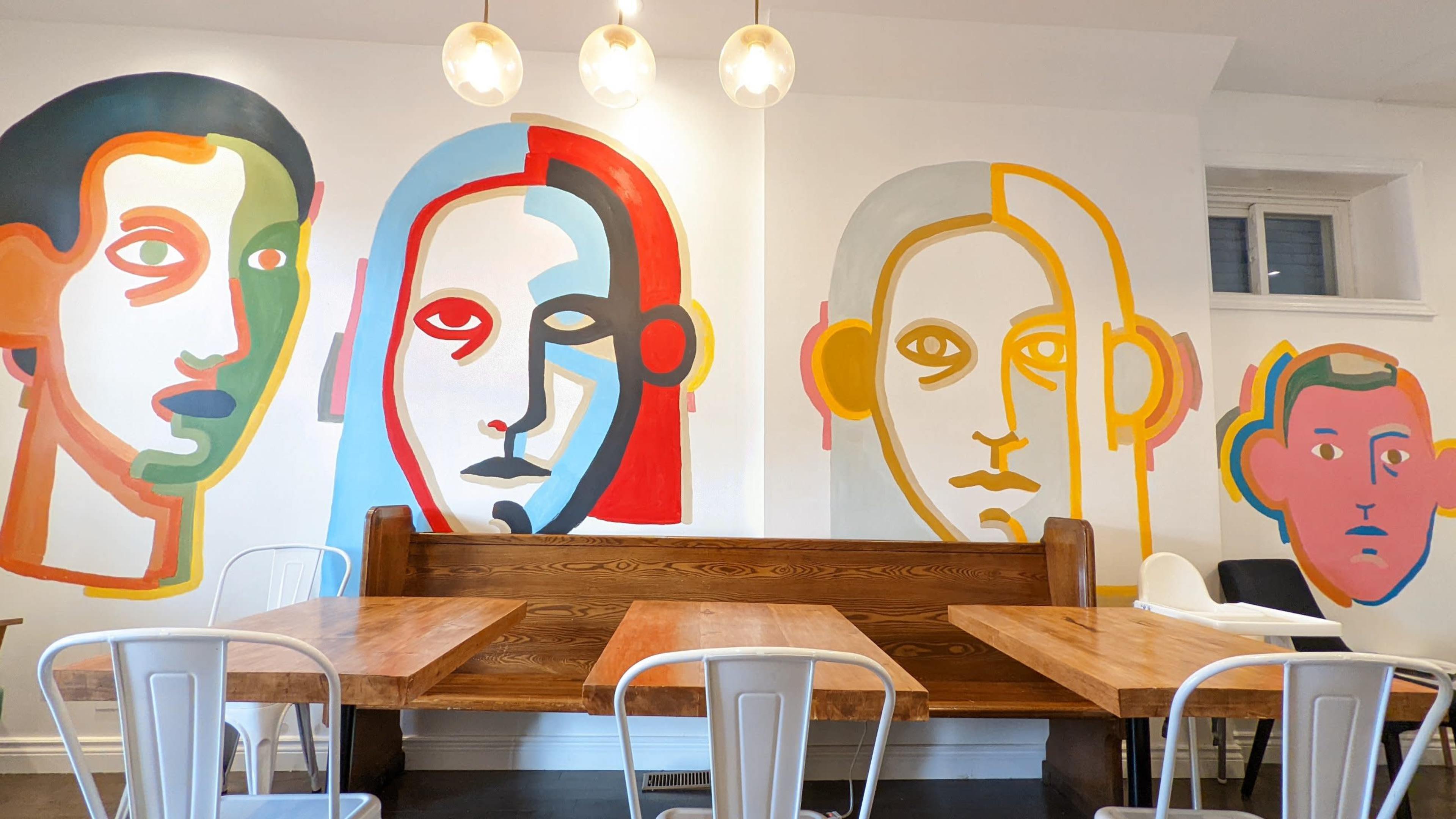 A café interior with wooden tables and chairs, illuminated by pendant lights, and a wall adorned with colorful abstract portraits.