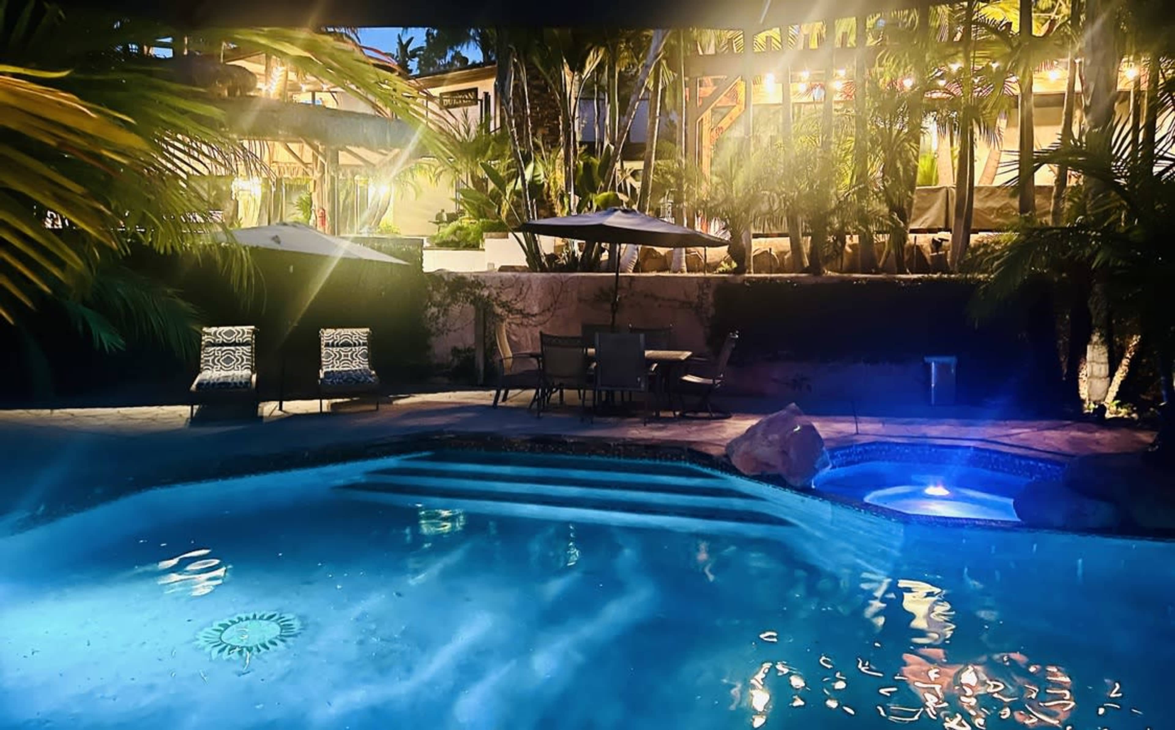 A serene backyard pool area features a lit pool, lounge chairs, and a patio surrounded by tropical plants and trees at dusk.