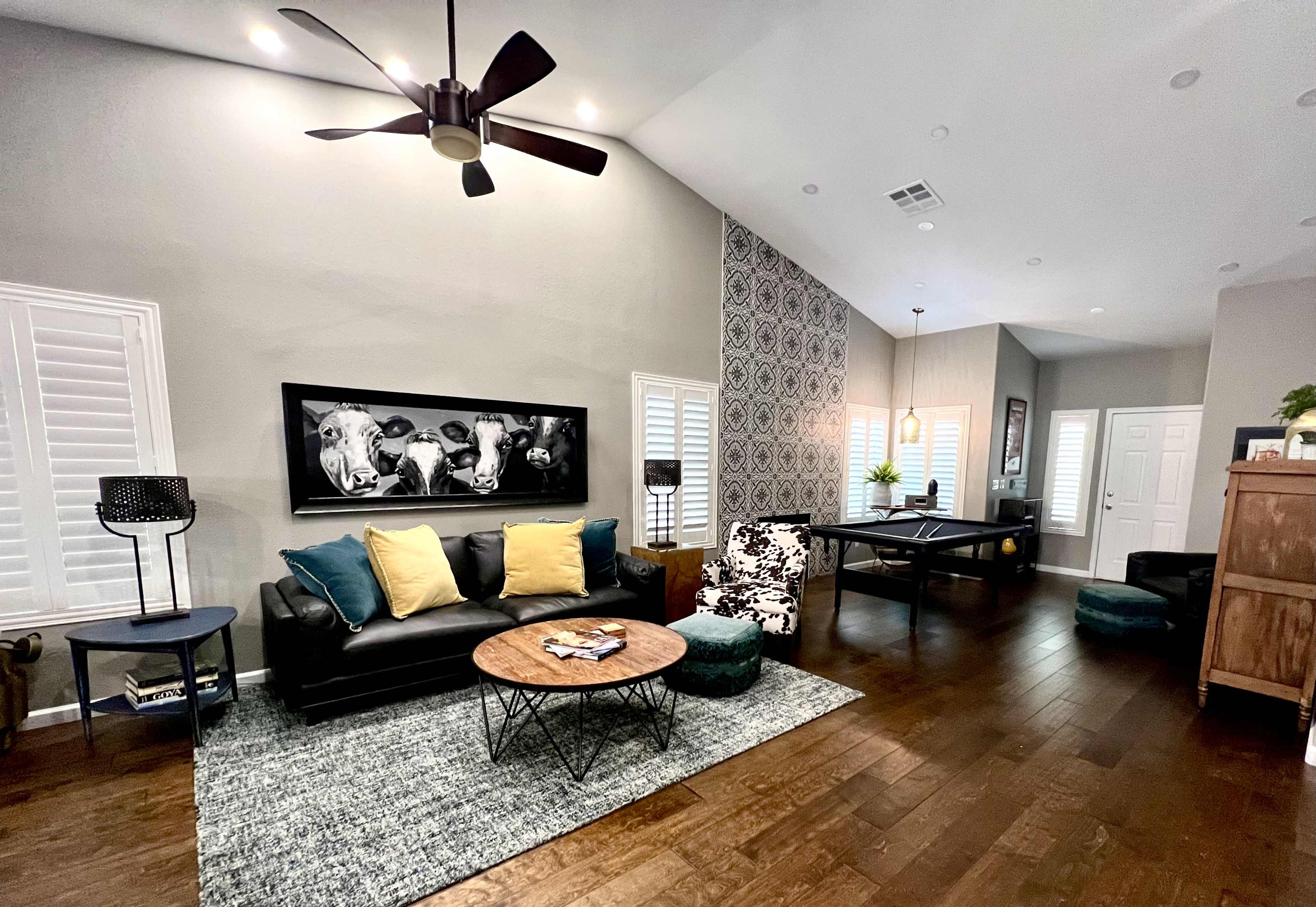 A modern living room features a black leather sofa with yellow cushions, a round coffee table, and a billiard table in the background.