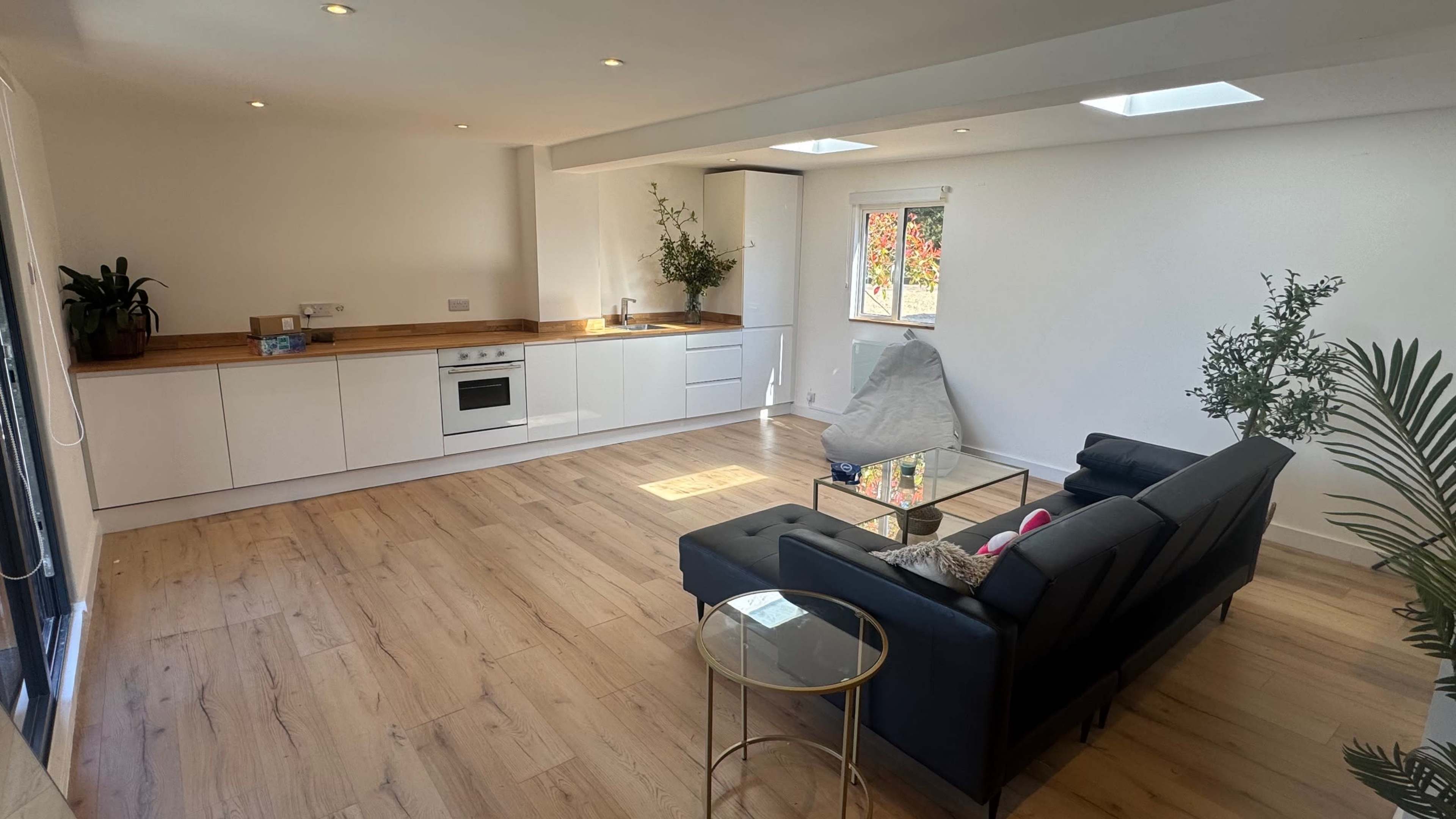 The image shows a modern living space with a kitchen area, a black sofa, a glass coffee table, and large windows that let in natural light.