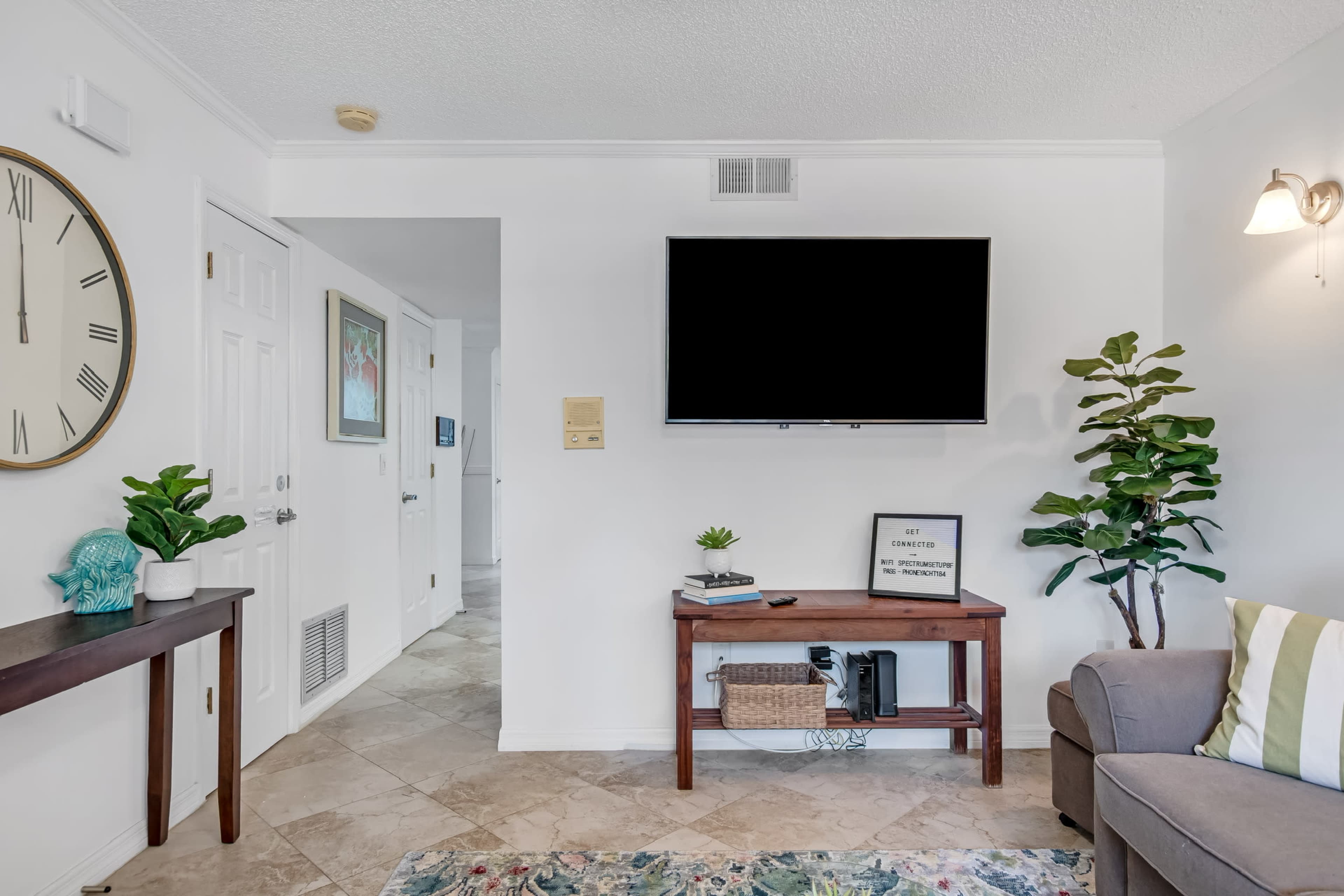 A living room with a wall-mounted television, a wooden table with decorative items, a green plant, and a light-colored couch.