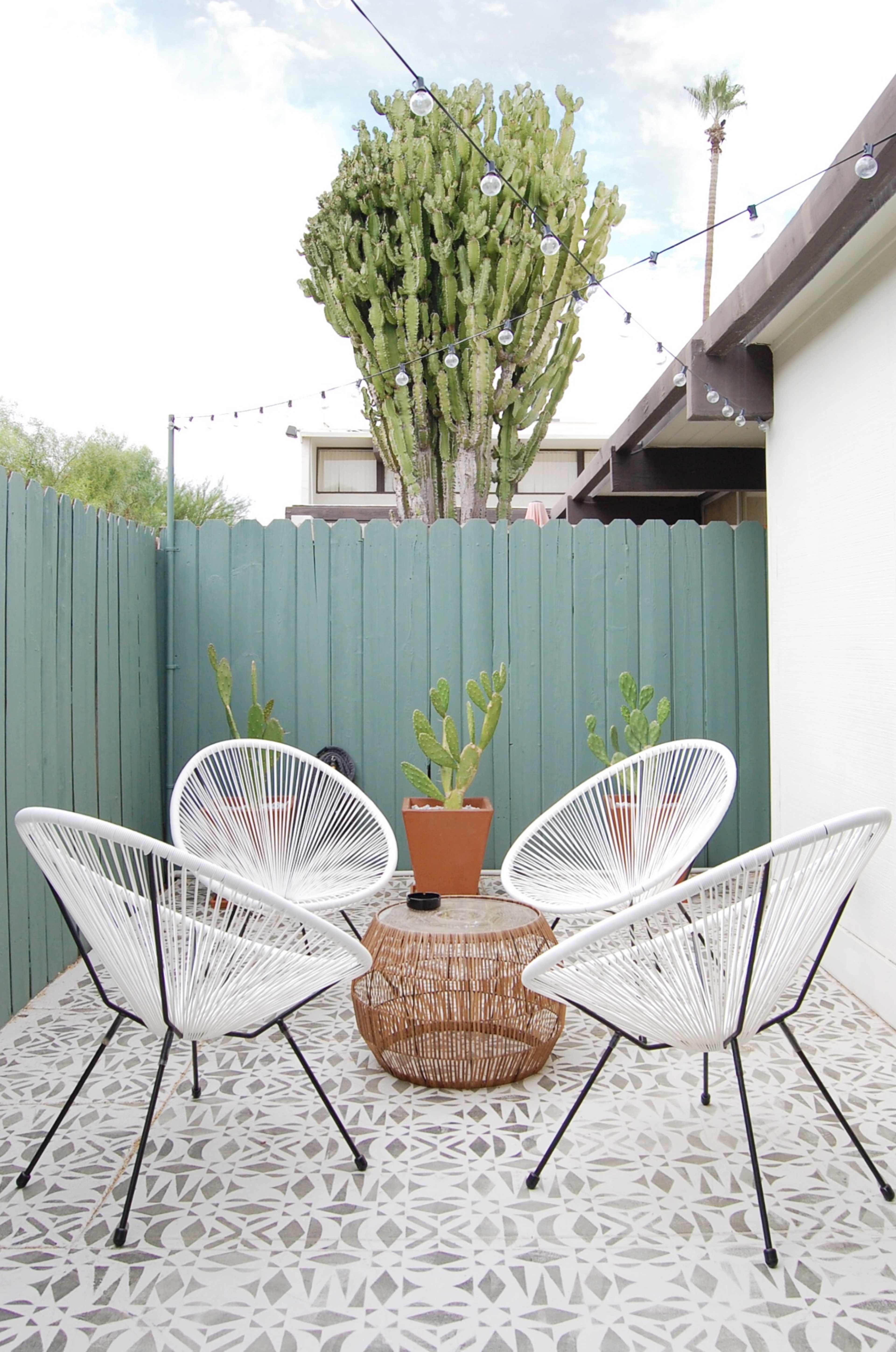 A patio features four white chairs arranged around a round wicker table, with a potted cactus in the center and a green wooden fence in the background.