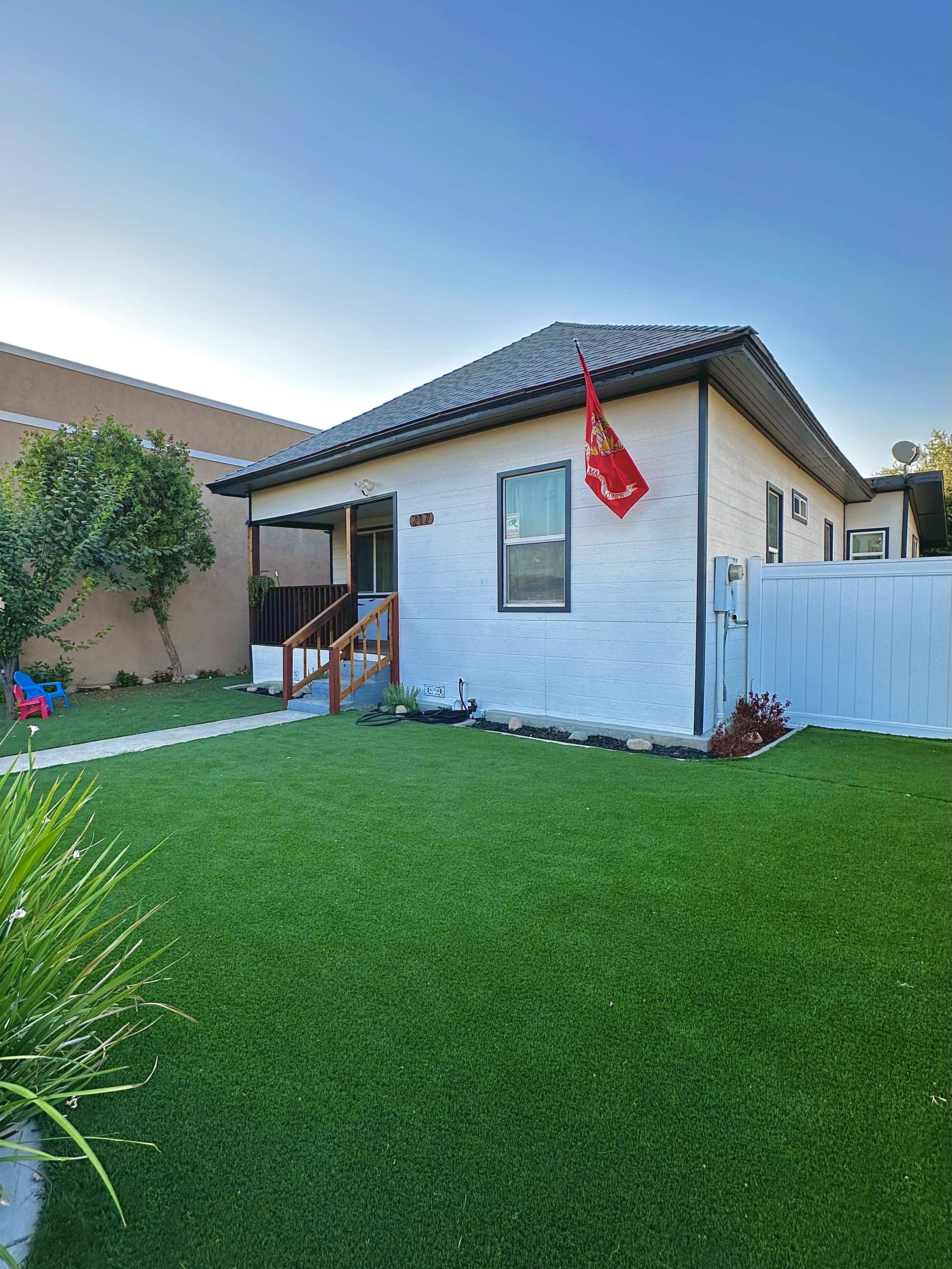 A small single-story house with a red flag displayed in front is surrounded by neatly trimmed green lawn and landscaping.