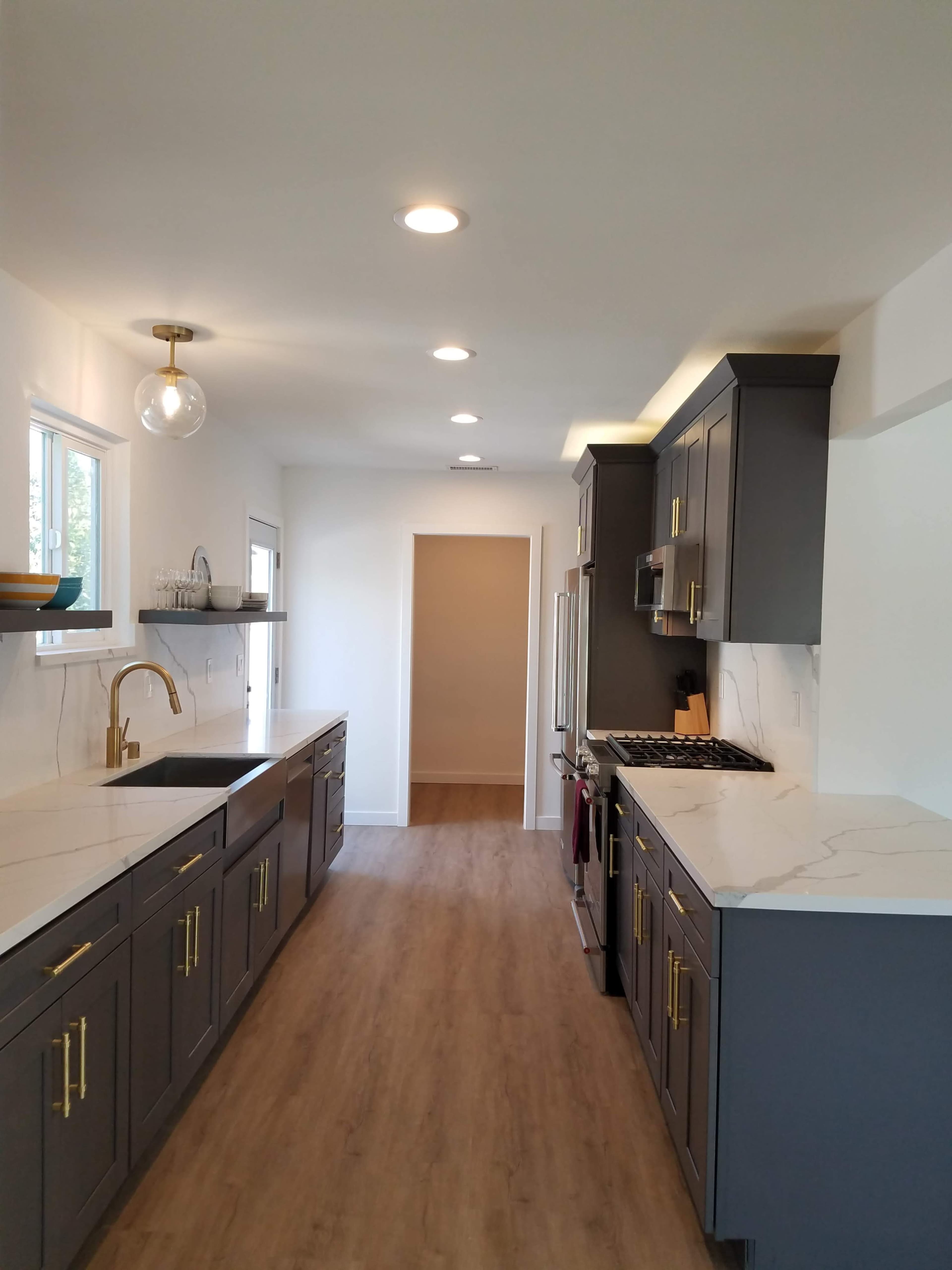 The image shows a modern kitchen featuring gray cabinets, marble countertops, and recessed lighting, with a view toward an adjacent hallway.