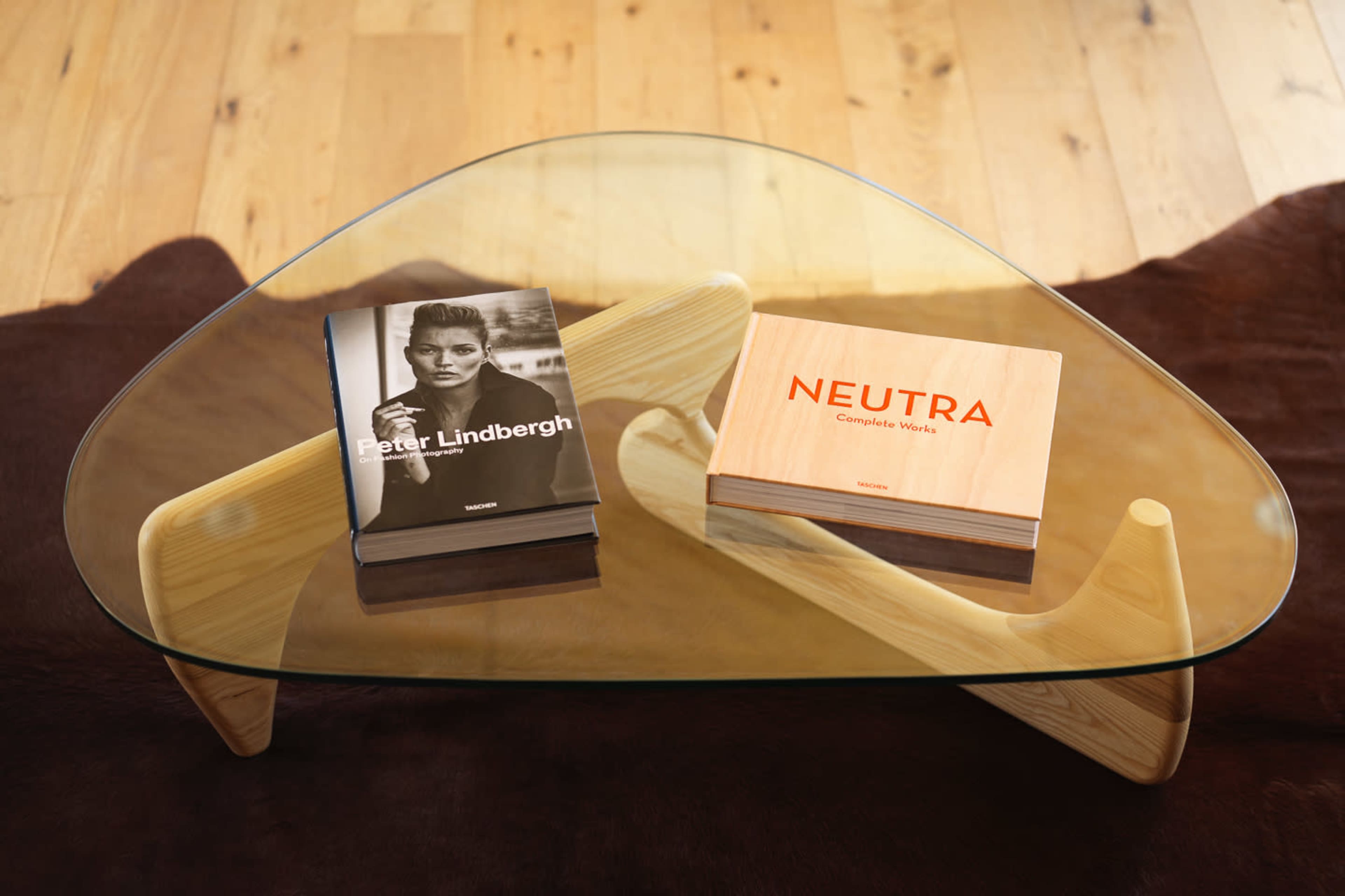 A glass coffee table with a wooden base displays two books: one titled "Peter Lindbergh" and the other "NEUTRA: Complete Works."