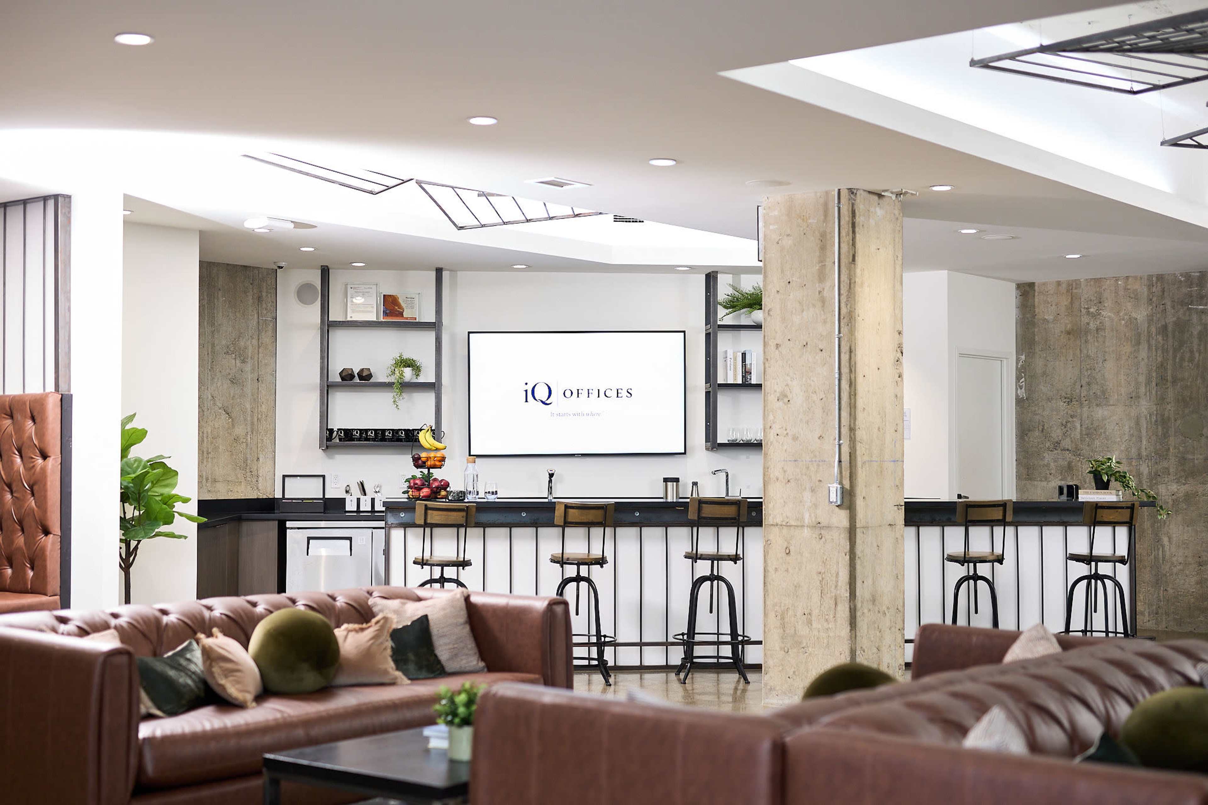 The image shows a modern office lounge area featuring a bar with seating, a television screen displaying the logo of iQ Offices, and a mix of comfortable seating arrangements.