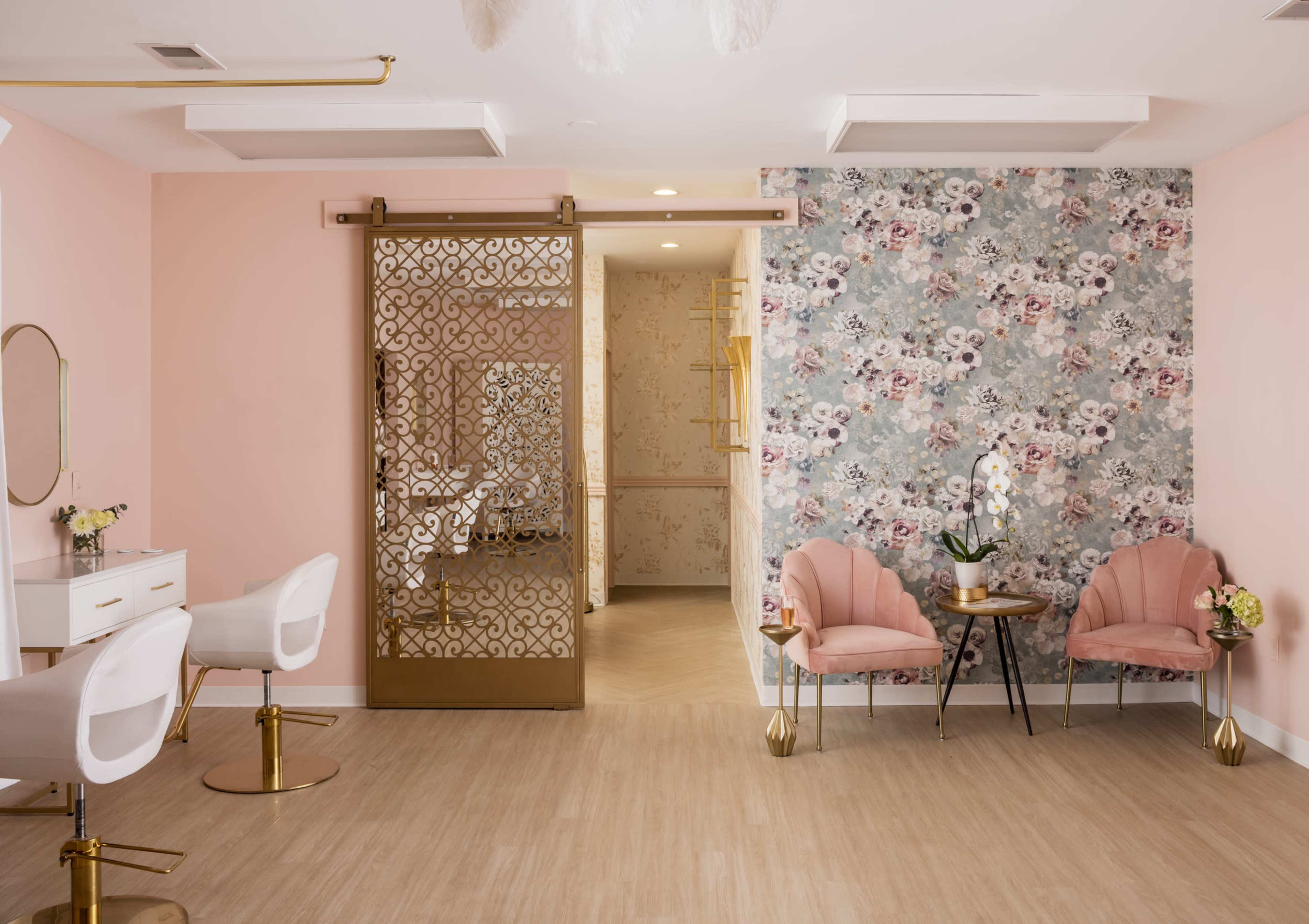 A stylish salon interior featuring gold accents, pink chairs, a decorative partition, and floral wallpaper.