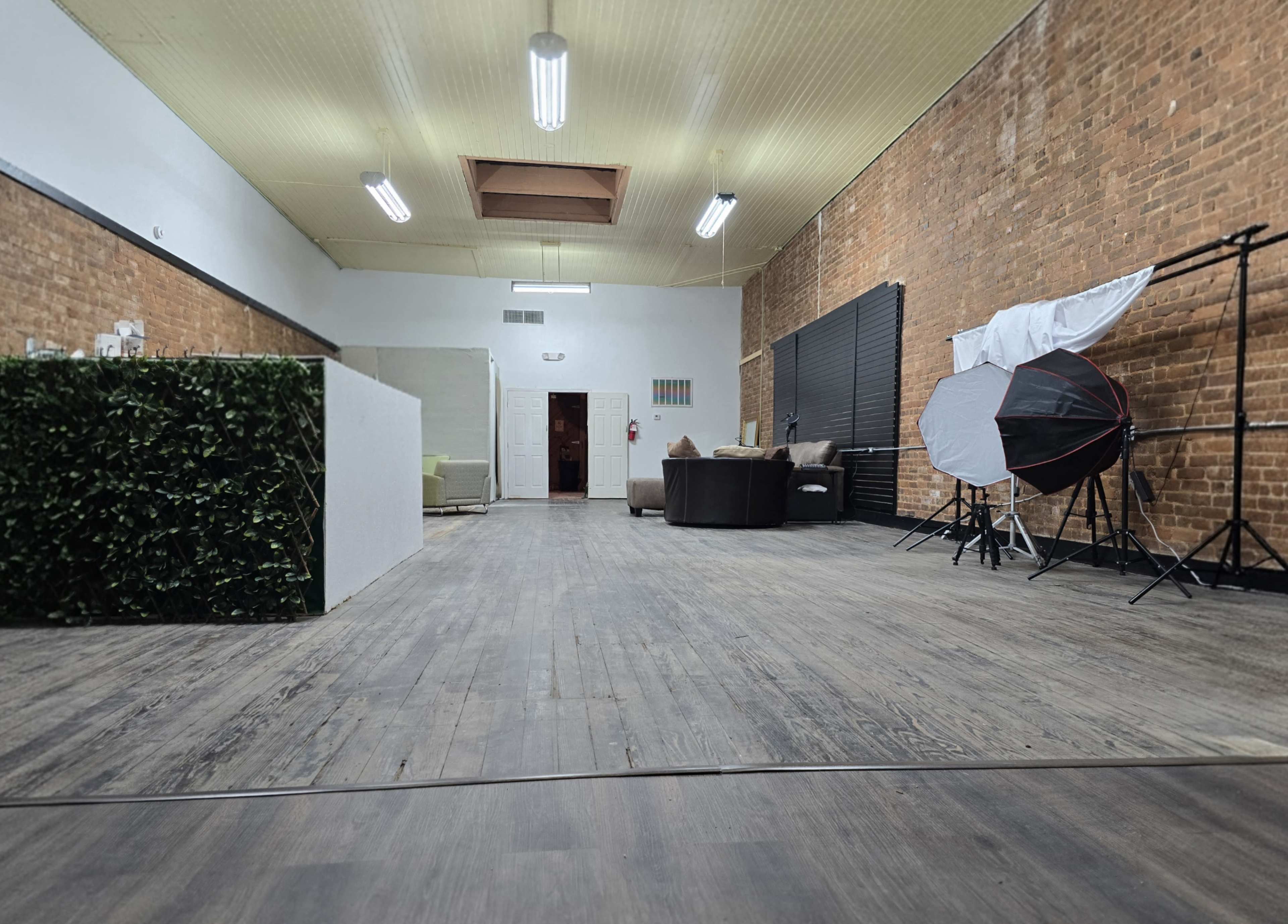The image shows a spacious, well-lit interior featuring exposed brick walls, wooden flooring, and furniture arranged in a modern layout.