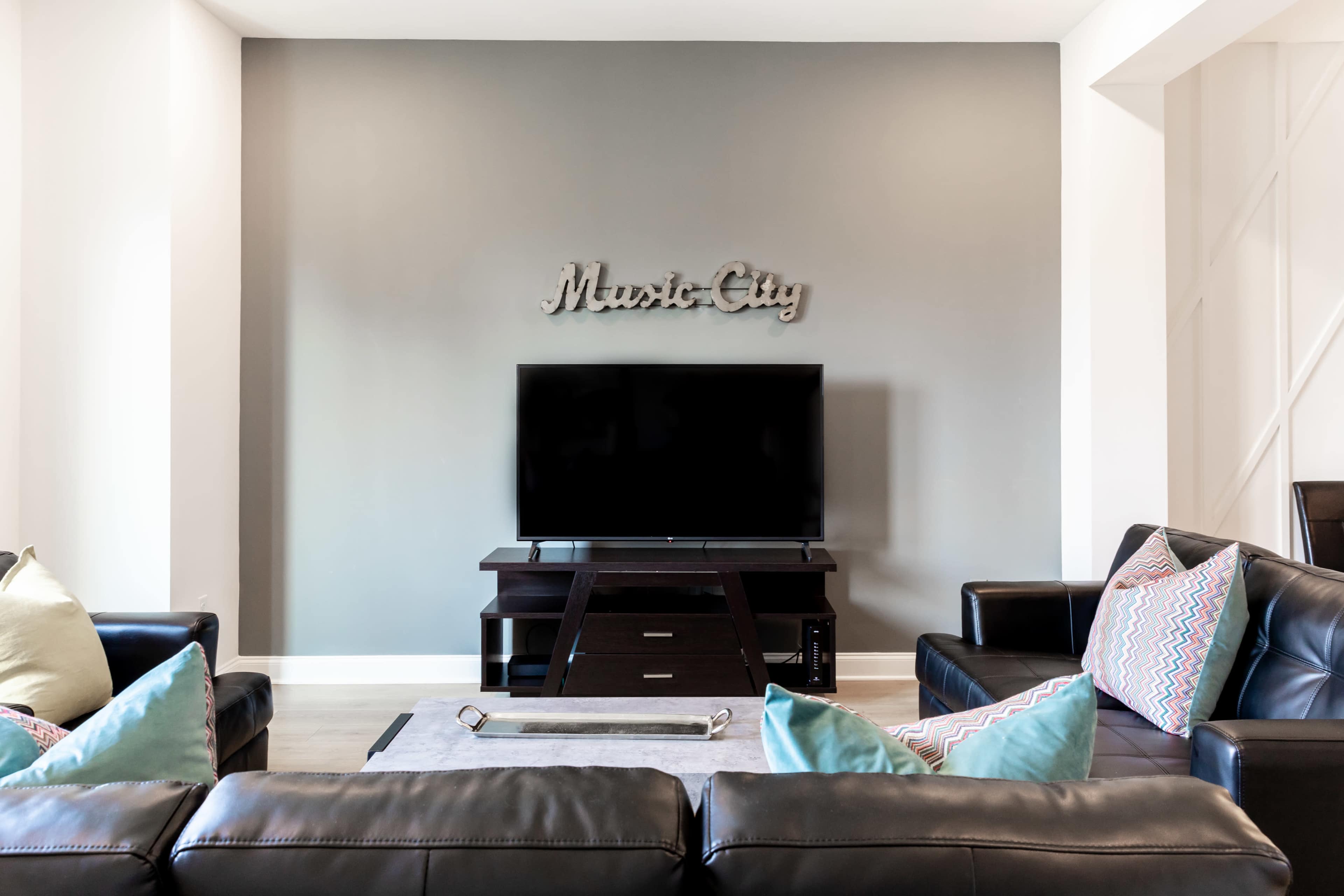 The image shows a living room with a black television mounted on a light gray wall, flanked by black leather sofas and decorative pillows.