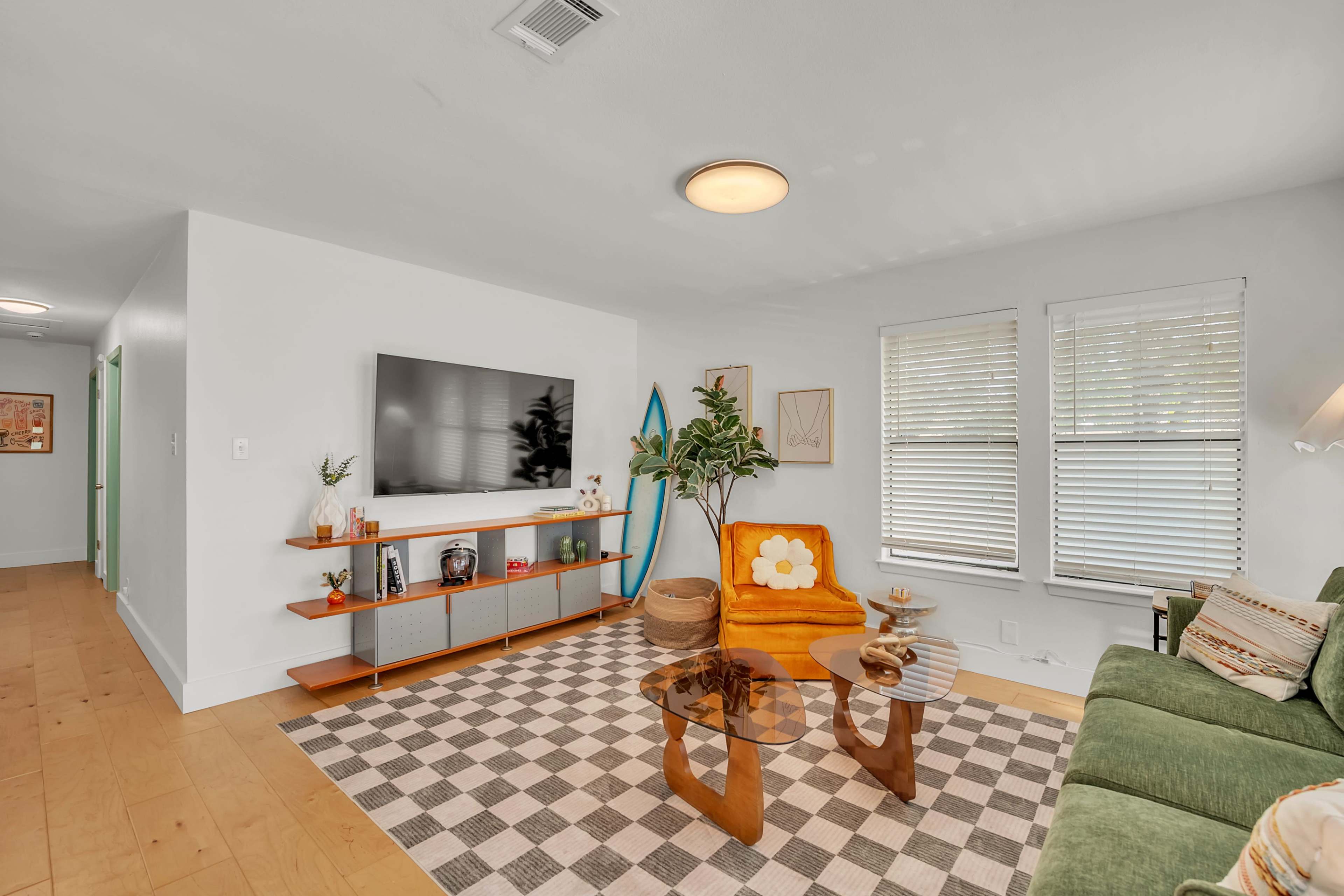 The living room features a colorful sofa, a wall-mounted television, and a coffee table on a patterned rug, with large windows allowing natural light to enter.
