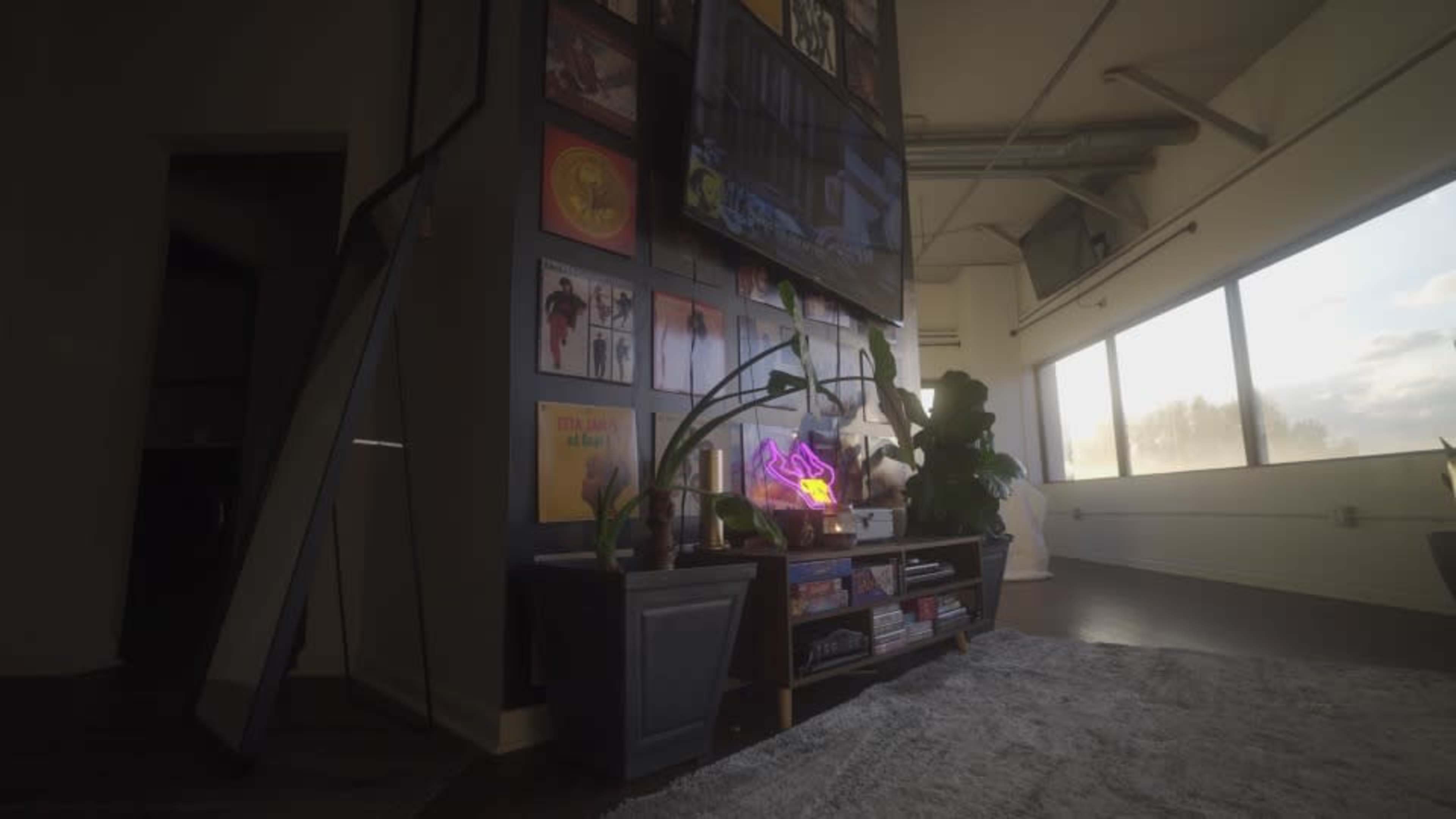 The image shows a modern living space with a dark wall featuring framed photographs, a shelf with decorative items, and a large window letting in natural light.