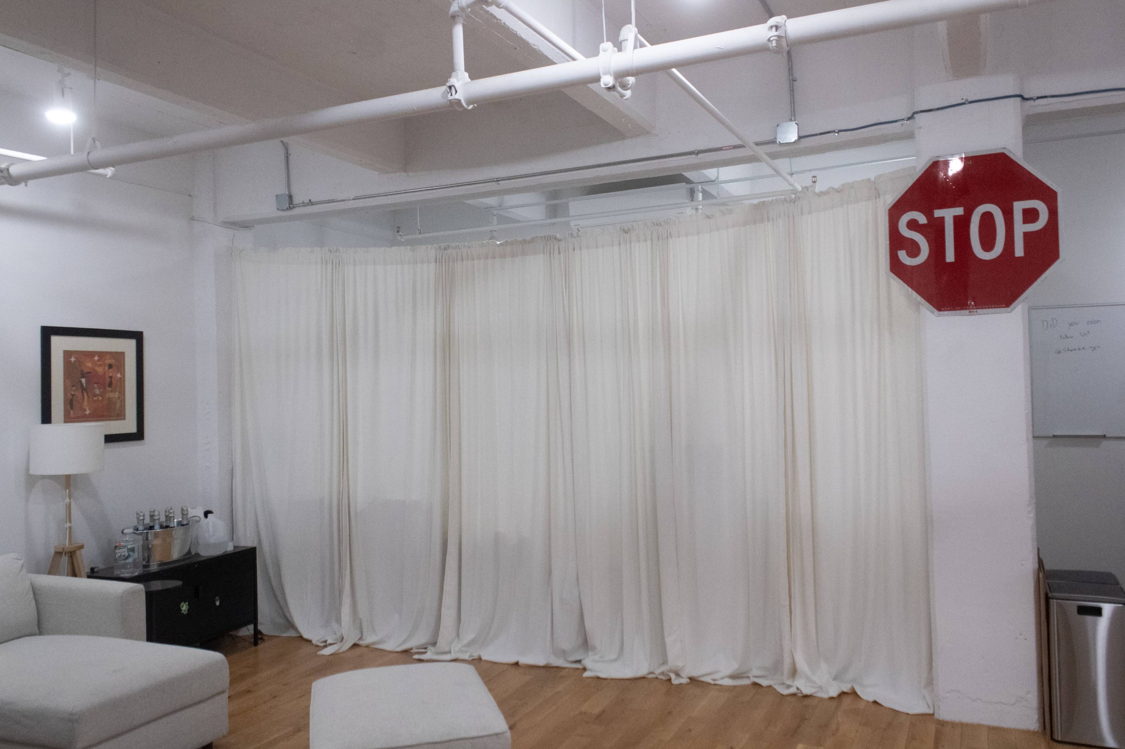 The image shows a soft-lit room featuring a white curtain backdrop, a stop sign mounted on the wall, a light-colored sofa, and a small black cabinet with bottles on top.