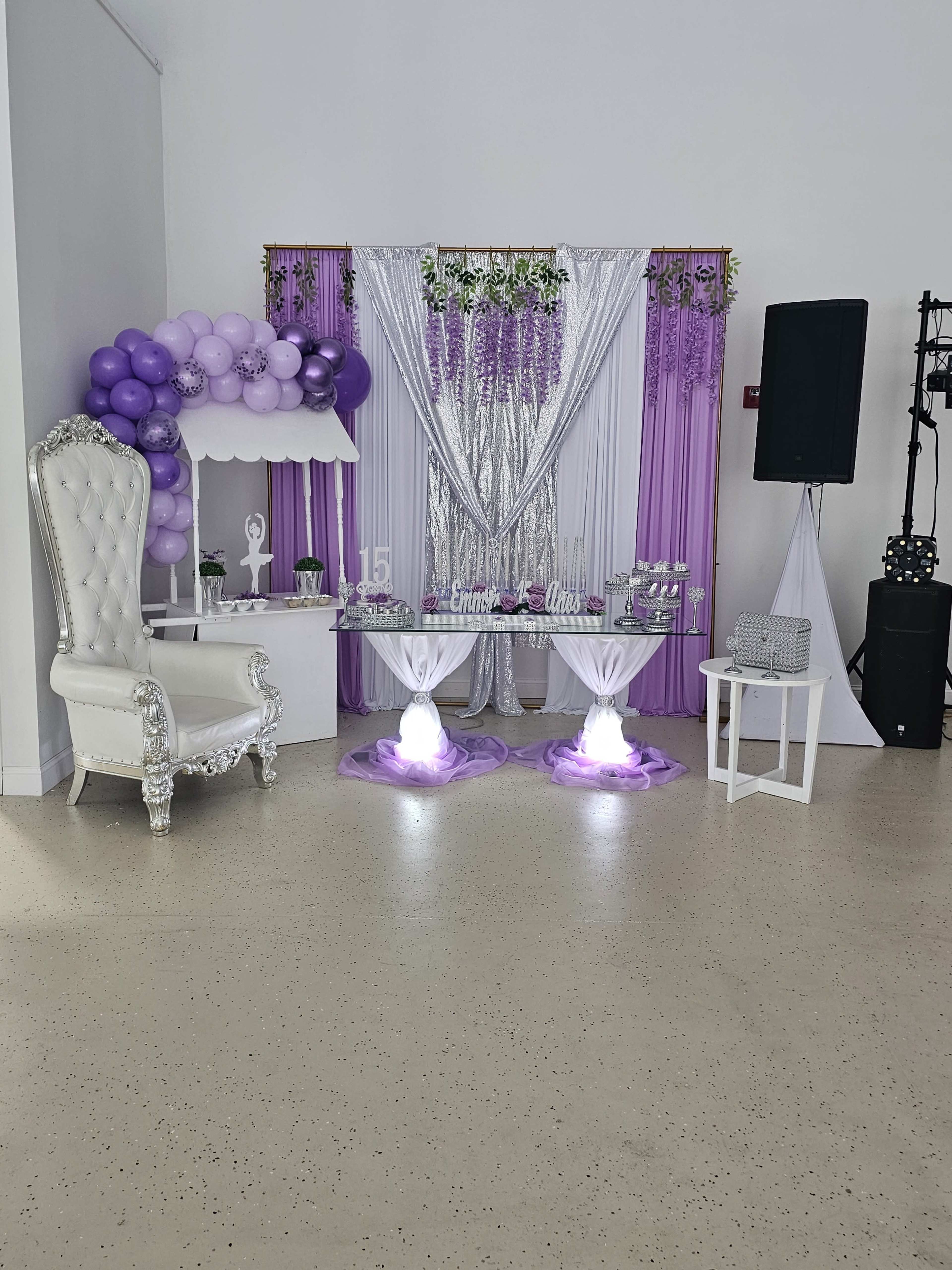 The image shows a decorated event space featuring a silver and purple theme, with a white throne chair, draped backdrops, and a dessert table illuminated from below.