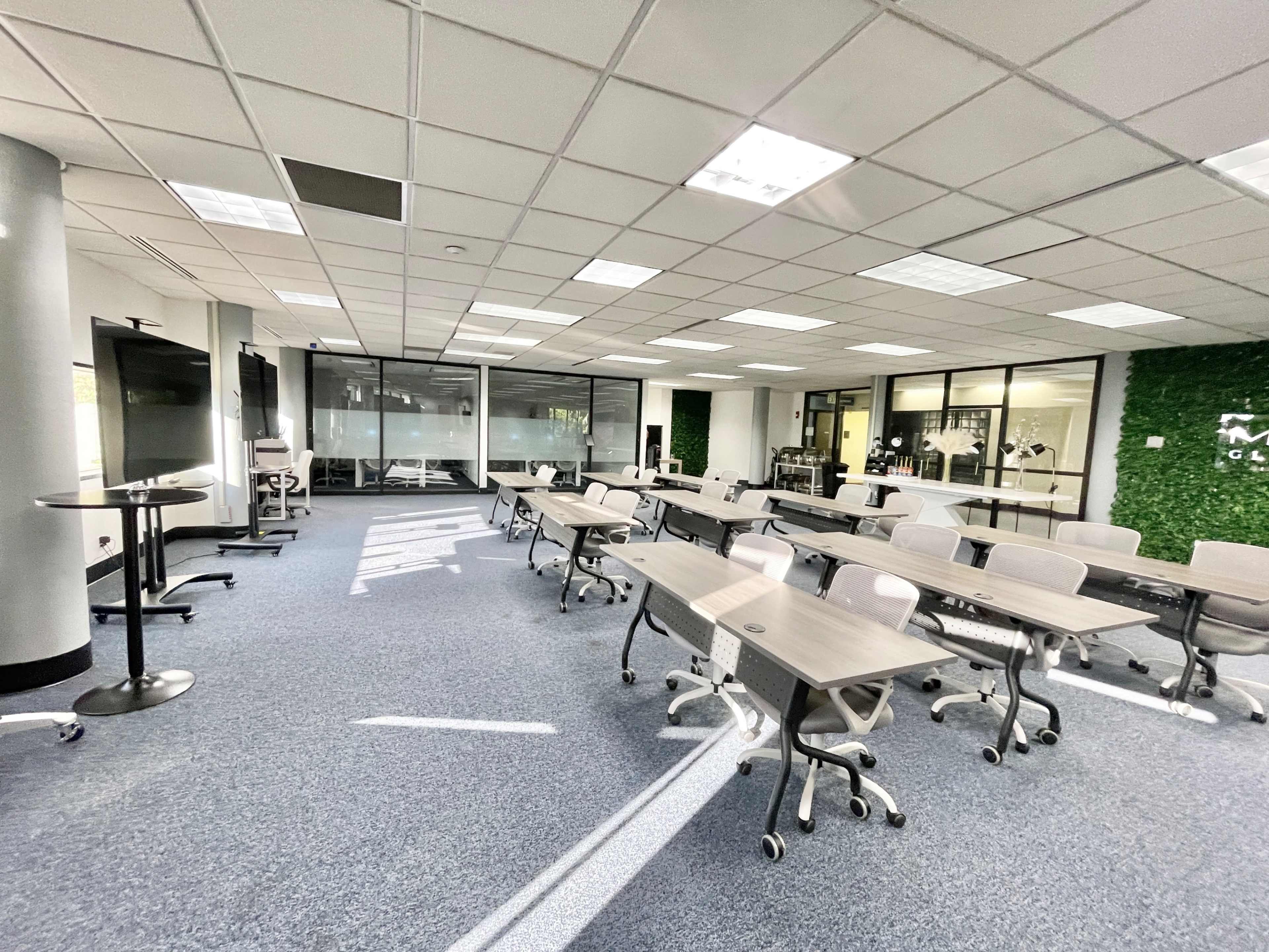 The image depicts a modern classroom setting with rows of movable tables and chairs, large windows allowing natural light, and a green wall feature in the background.