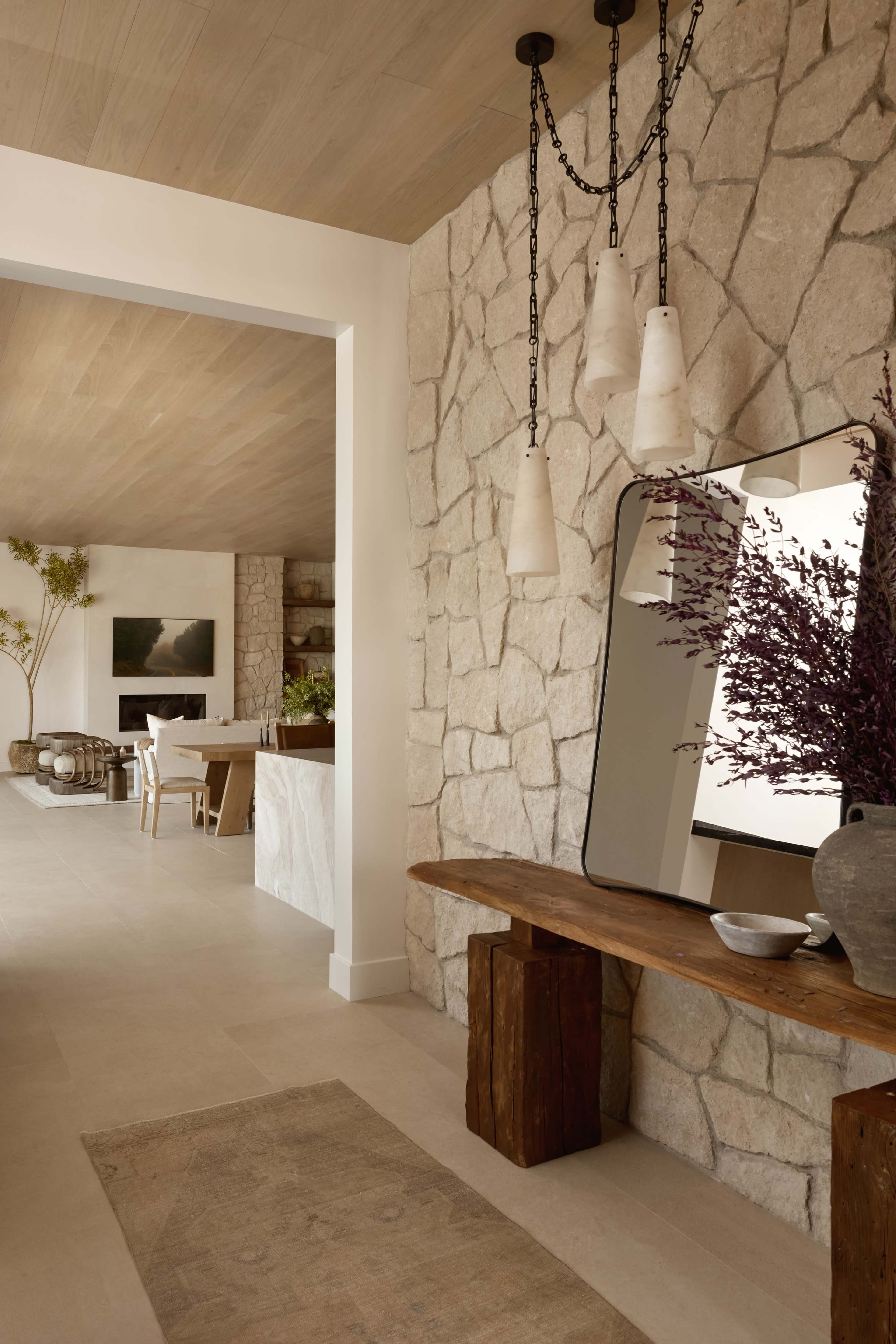 The image depicts a modern hallway with a stone wall, wooden bench, and decorative items, leading to an open living space.