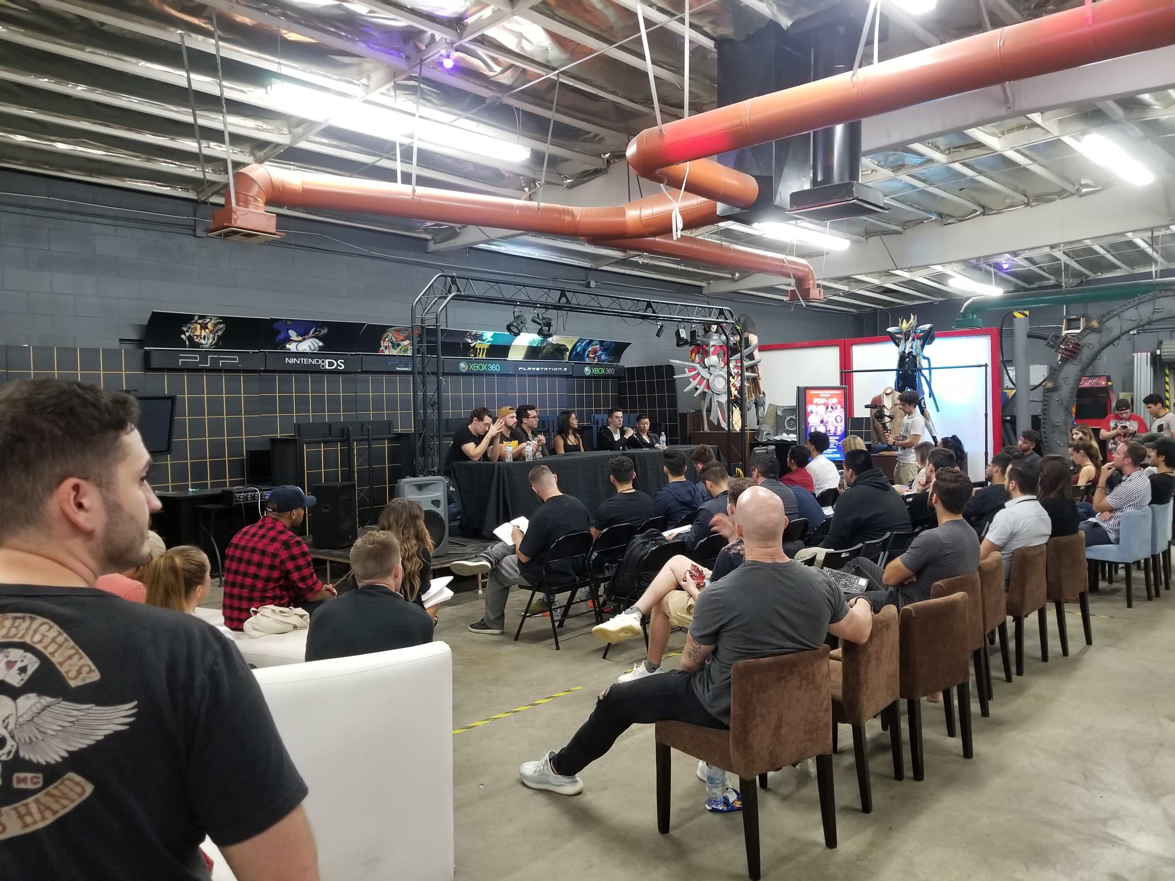 A group of speakers is seated at a long table facing an audience in a spacious indoor venue.