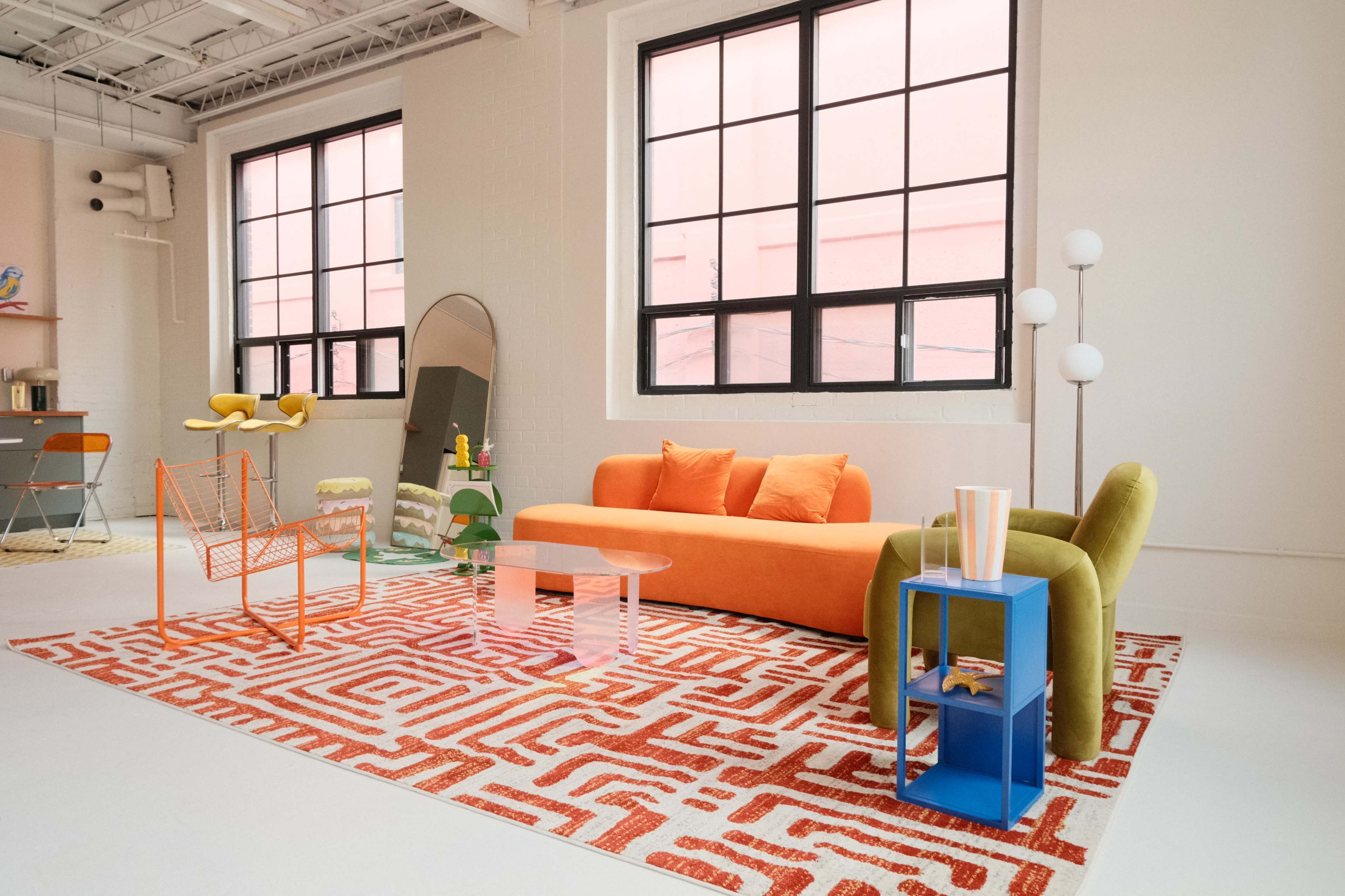 The image shows a brightly colored modern living room featuring an orange sofa, green armchair, patterned rug, and various decorative furniture pieces.