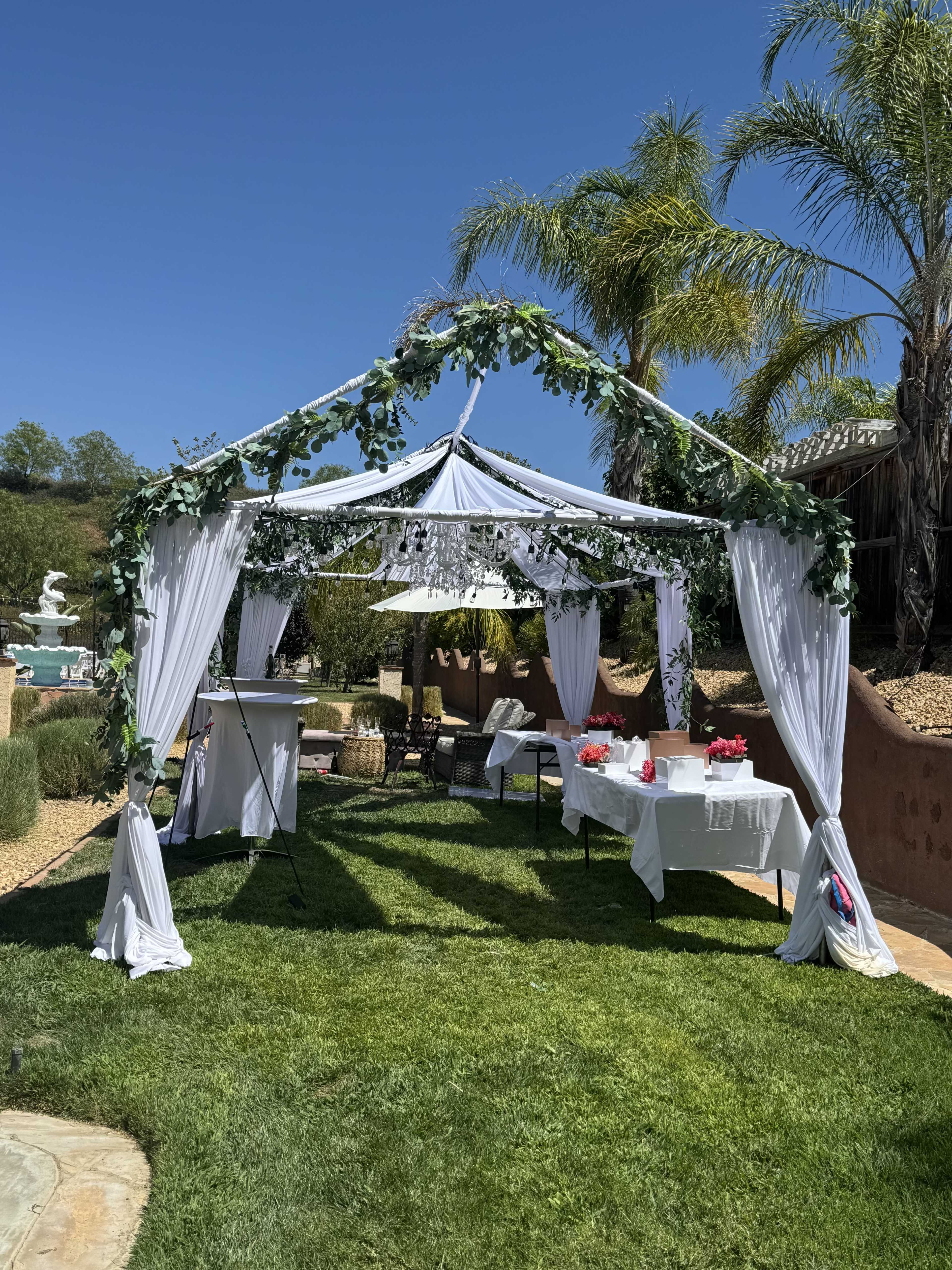 A white canopy adorned with greenery is set up in a grassy area, surrounded by palm trees and a decorative fountain in the background.