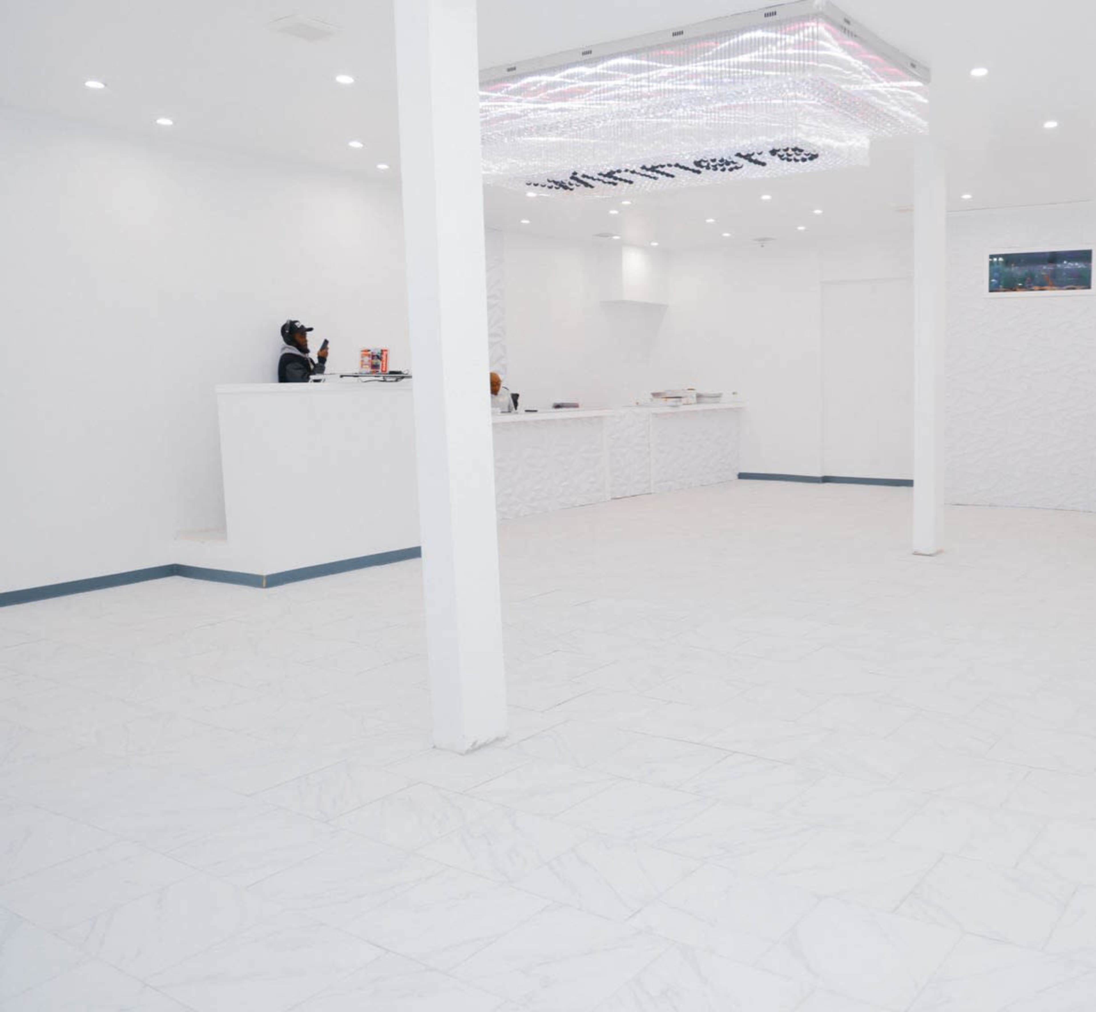 A spacious, well-lit interior features white walls and flooring, with a front desk and a staff member attending to an area behind the counter.