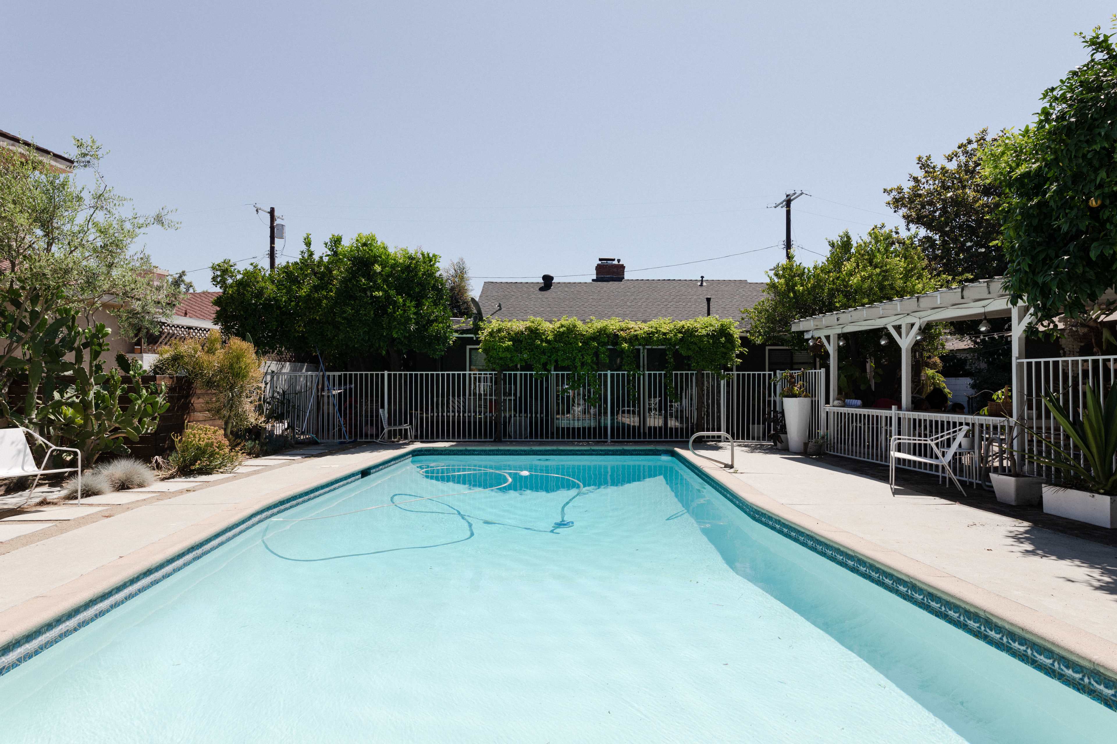 The image shows a clear swimming pool surrounded by a fenced area, with trees and shrubs in the background.