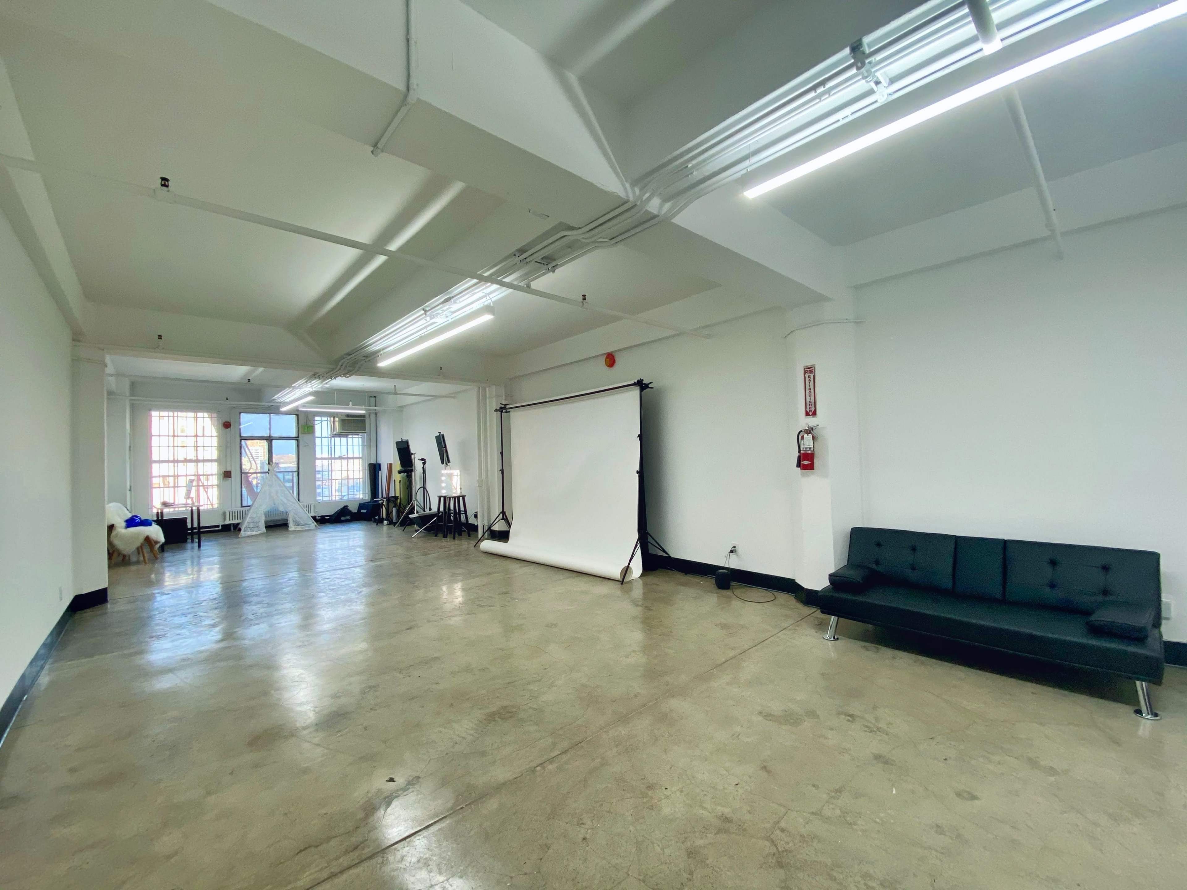 A spacious, empty room with concrete flooring and large windows, featuring a black sofa, a backdrop stand, and various studio equipment.