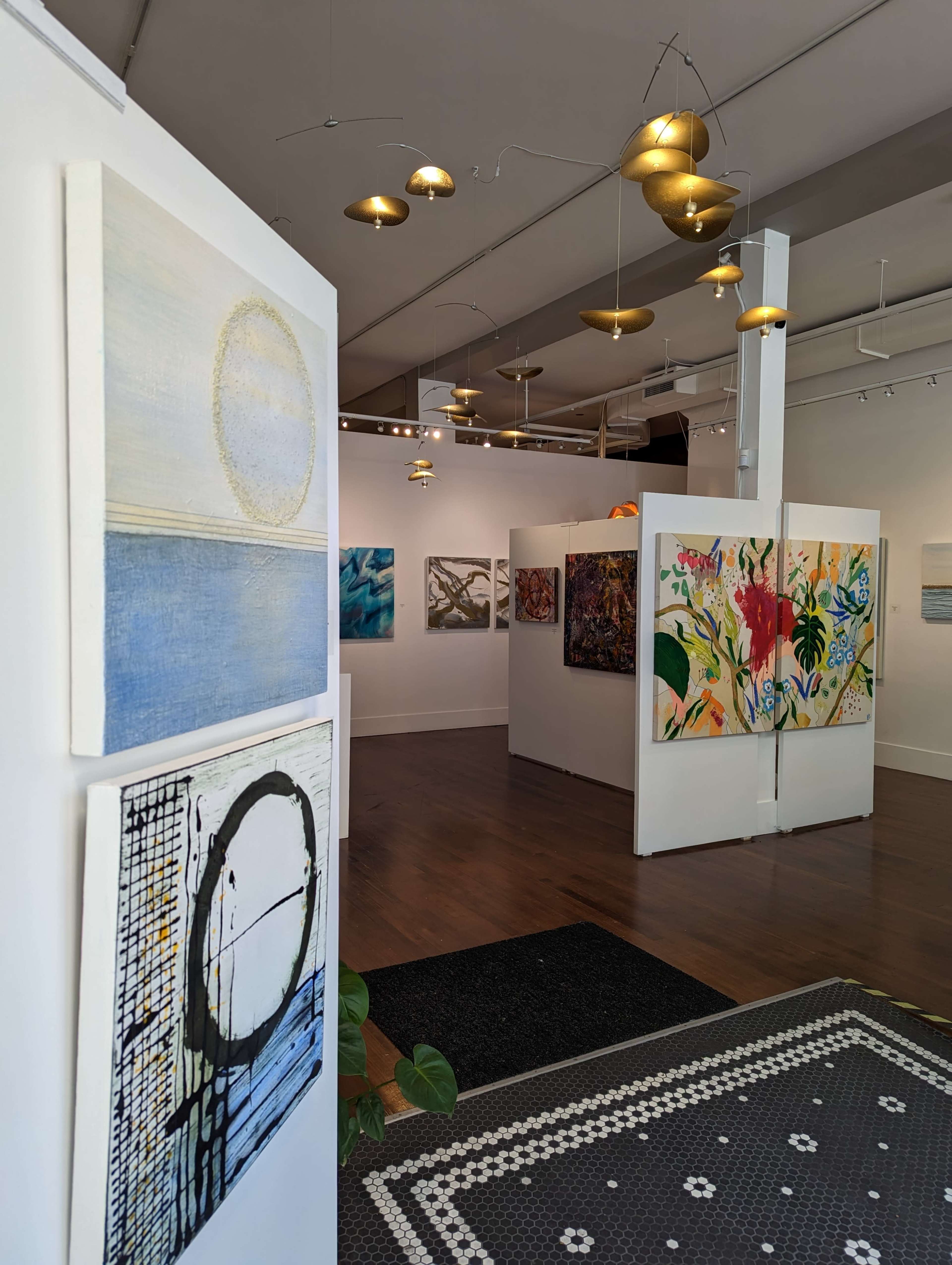 An art gallery interior featuring various paintings displayed on white walls, with a patterned rug at the entrance and modern lighting fixtures above.