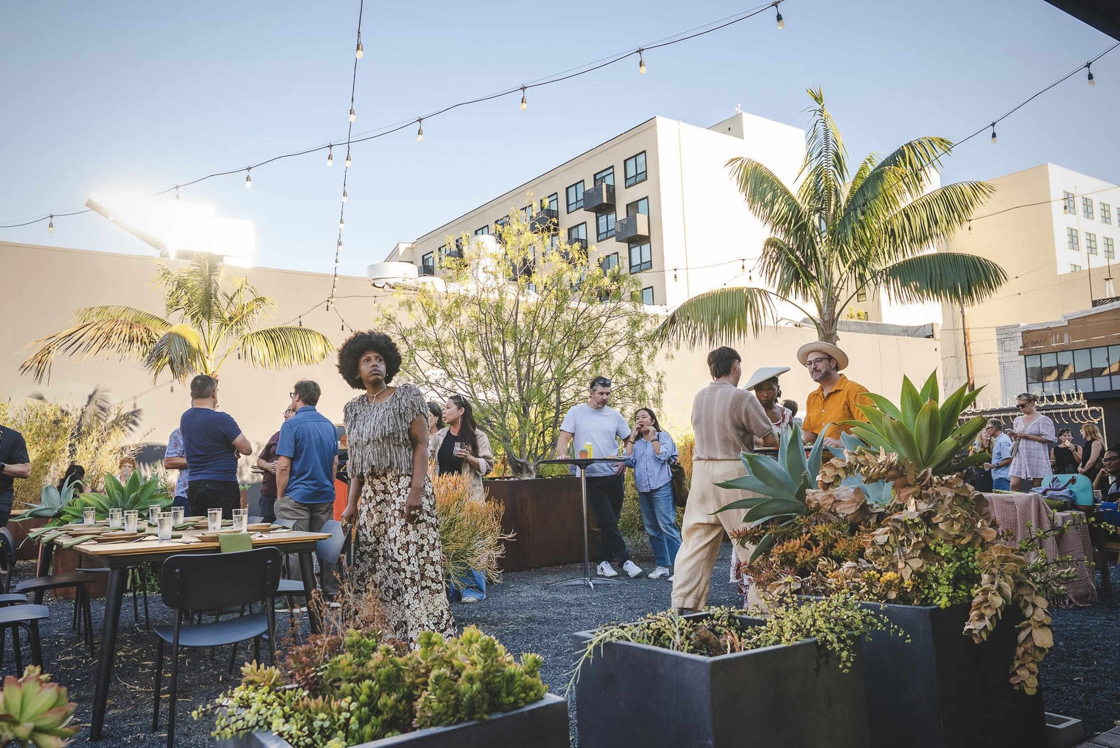 A lively outdoor event takes place on a rooftop, featuring guests mingling among tropical plants and string lights.