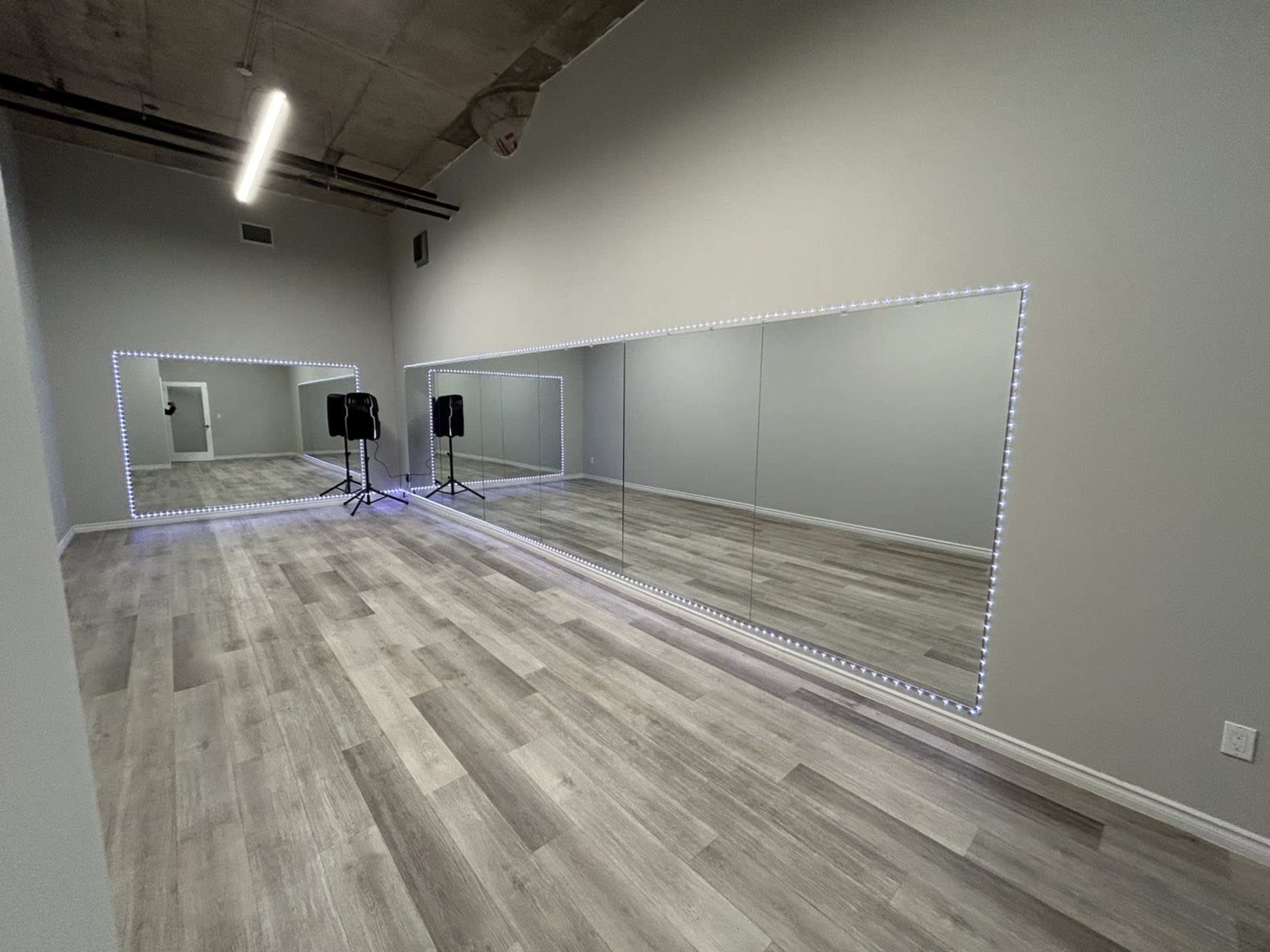 The image depicts a spacious room with large mirrors bordered by LED lights, wooden flooring, and two portable speakers positioned near the back wall.