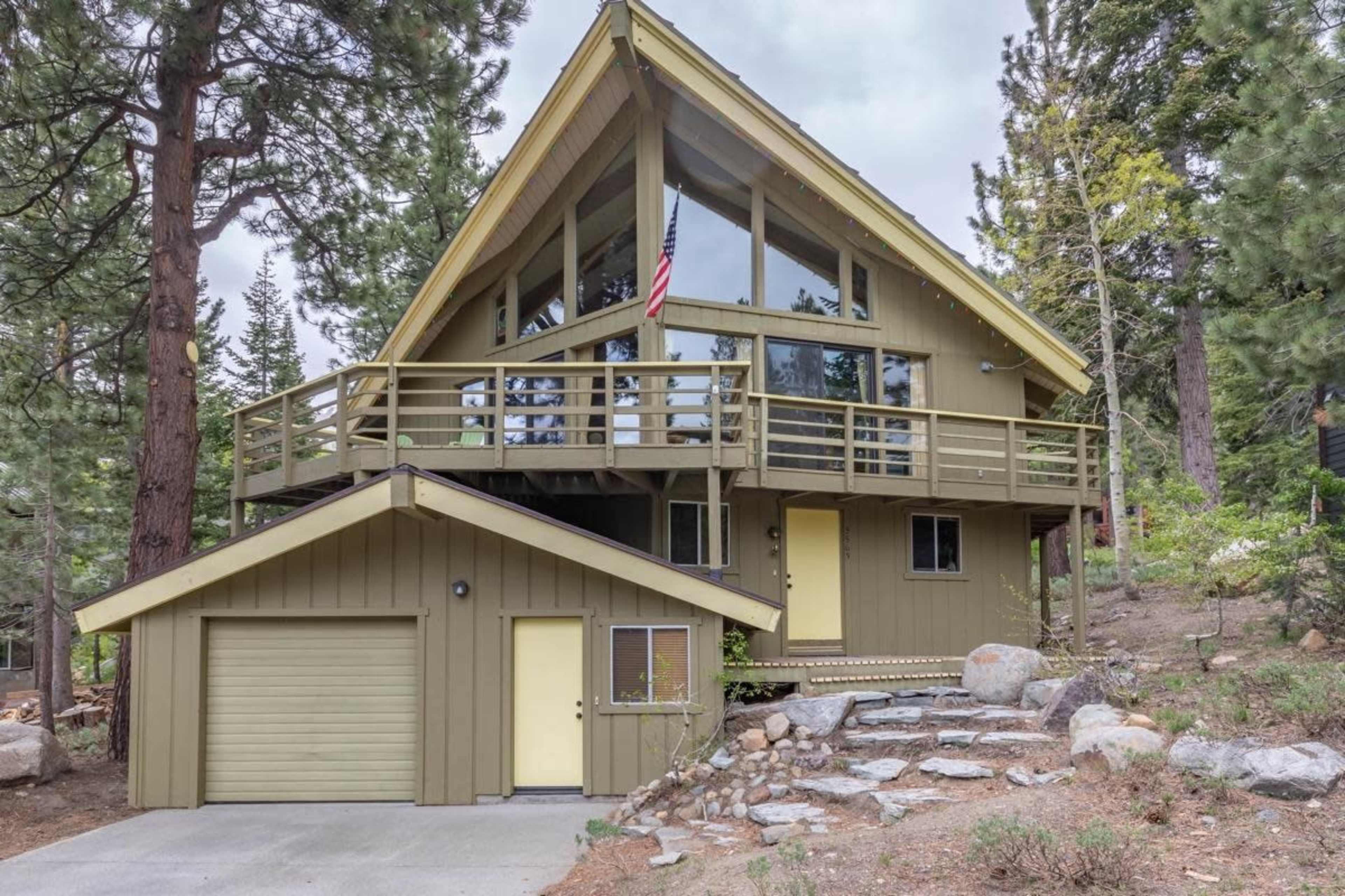 A two-story, A-frame house with large windows, a balcony, and a garage, situated among trees in a mountainous area.