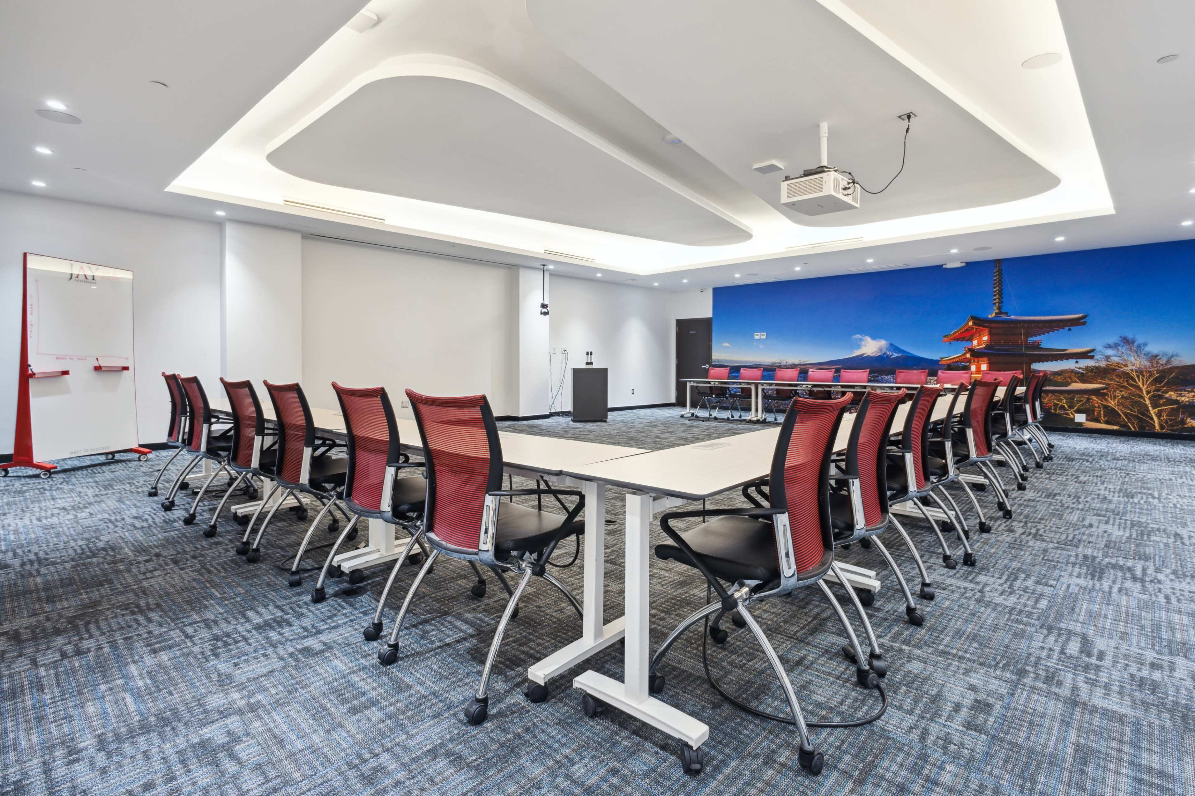 A conference room features rows of chairs arranged around tables, with a large screen displaying a scenic backdrop.