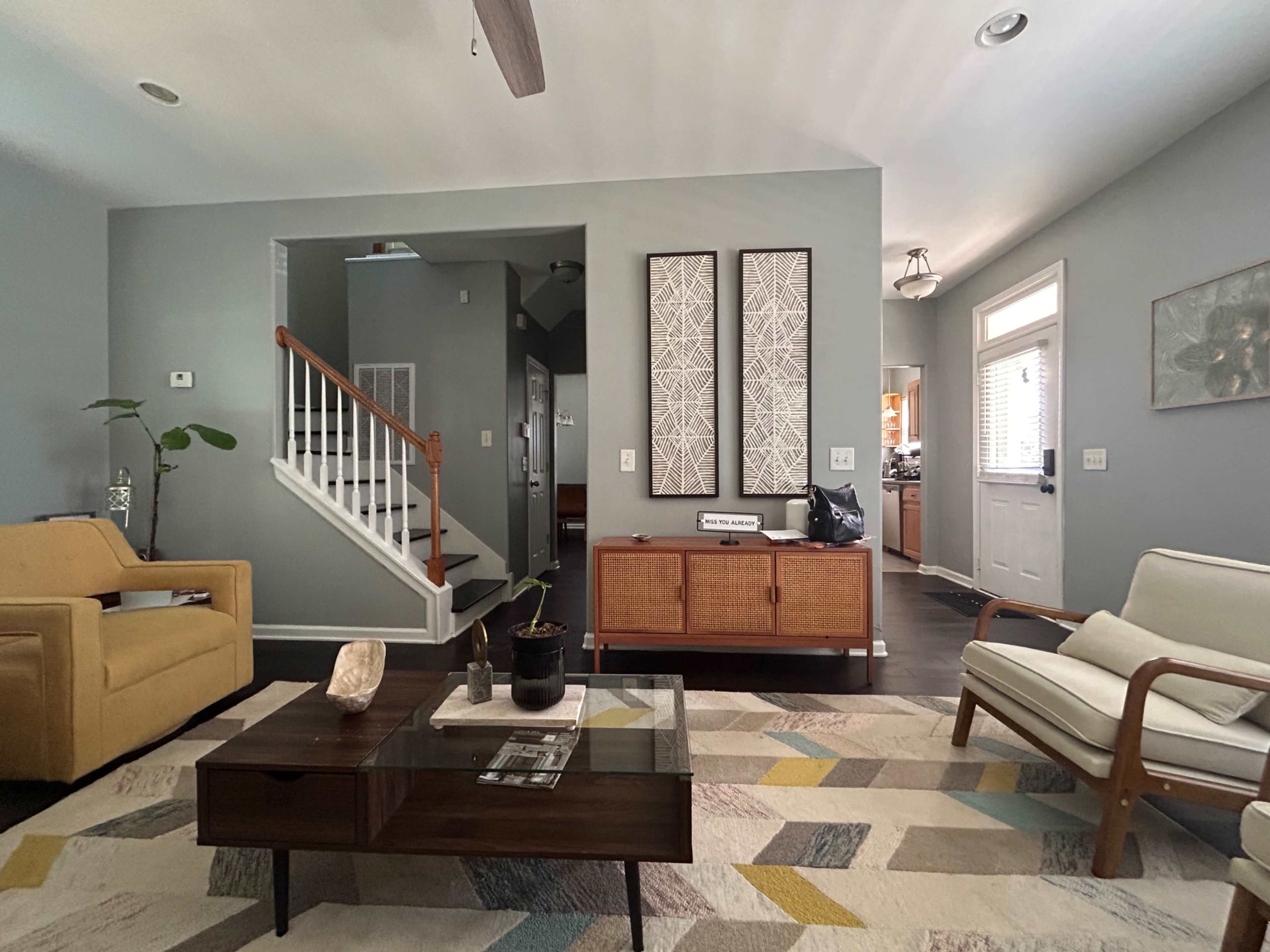 A living room features a yellow armchair, a white armchair, a wooden coffee table, and a staircase in the background.