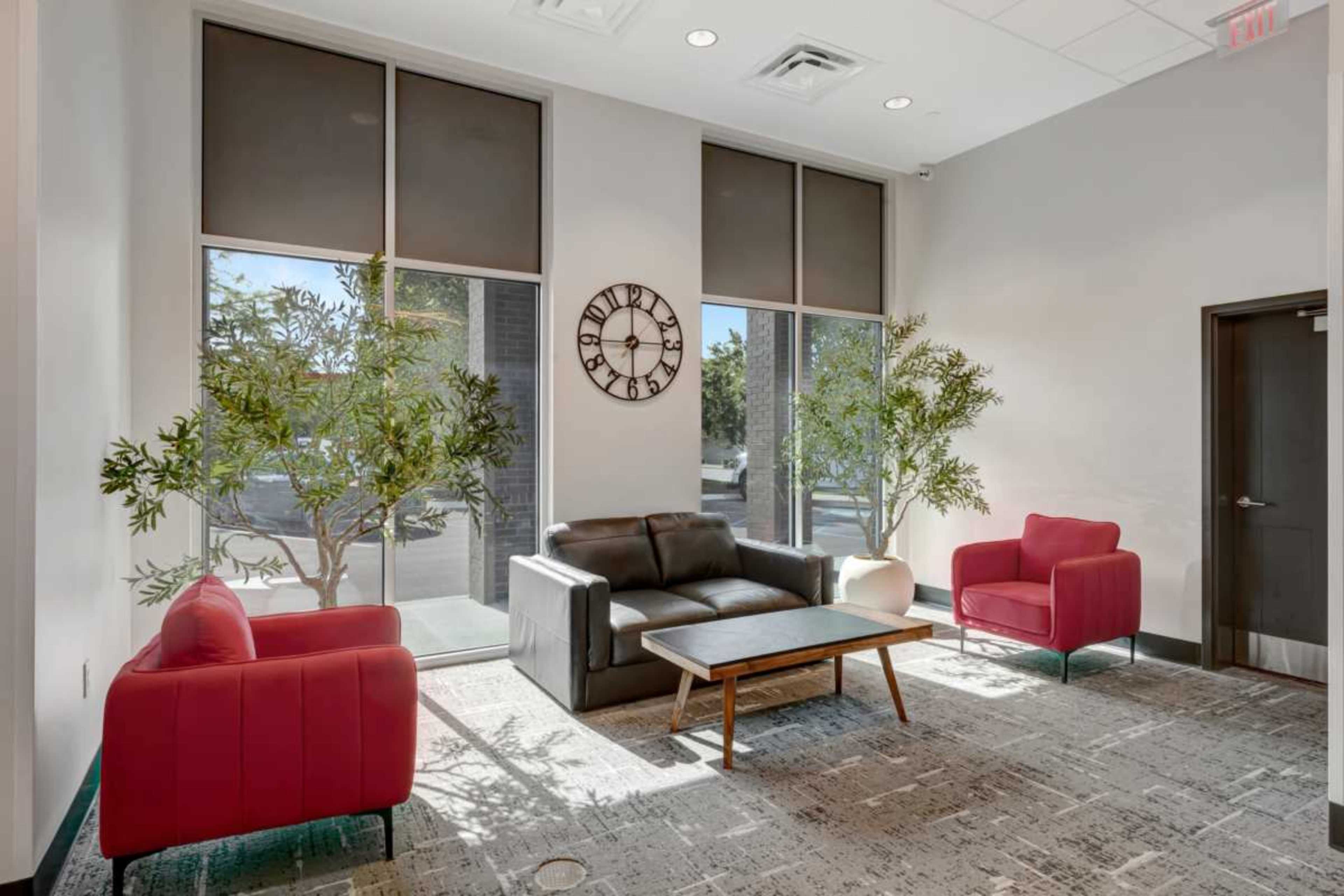 A modern seating area features a black leather sofa, two red chairs, a wooden coffee table, and two trees near large windows.