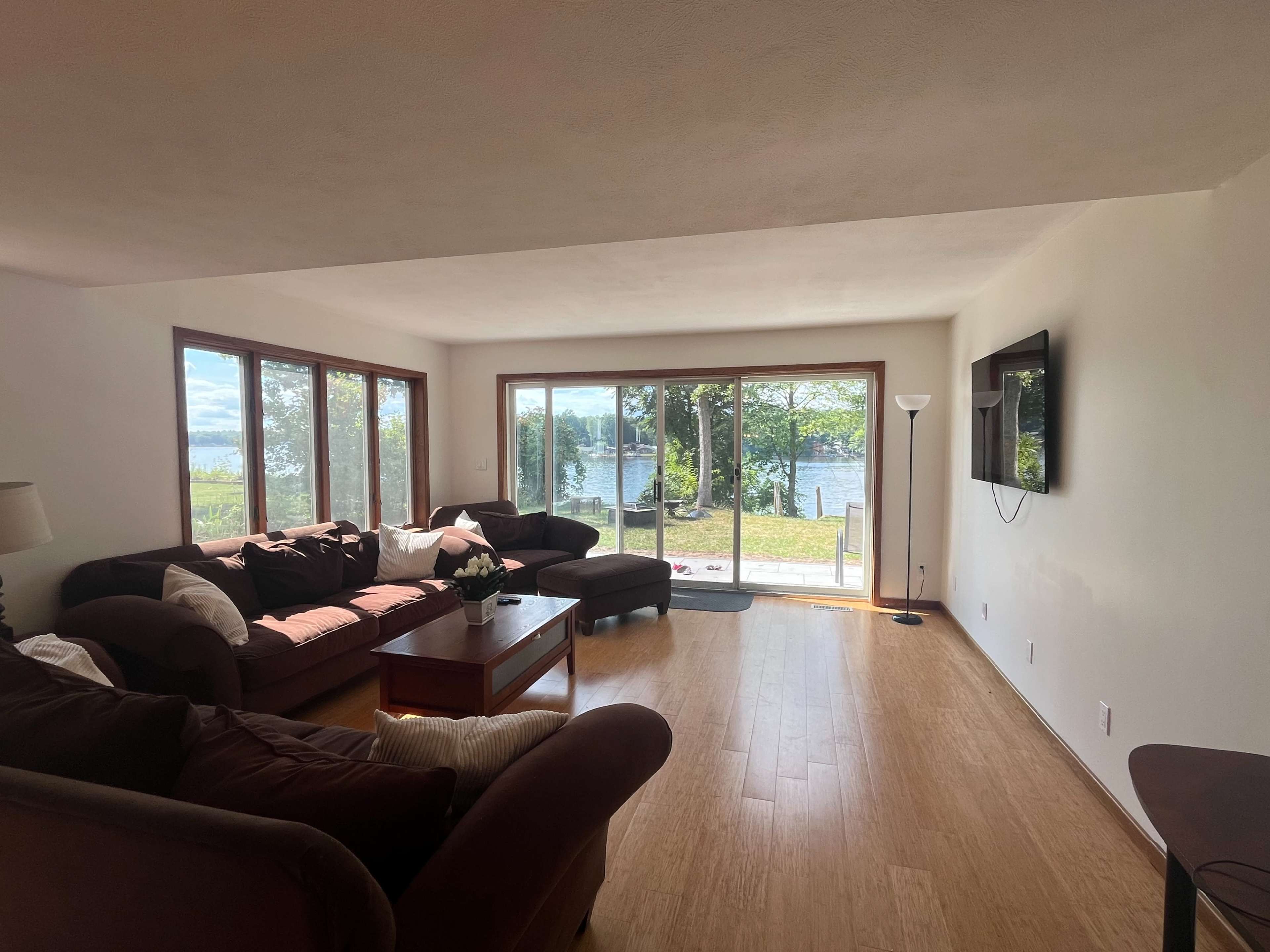 A spacious living room features large windows with a view of a lake, furnished with brown couches and a wooden coffee table.