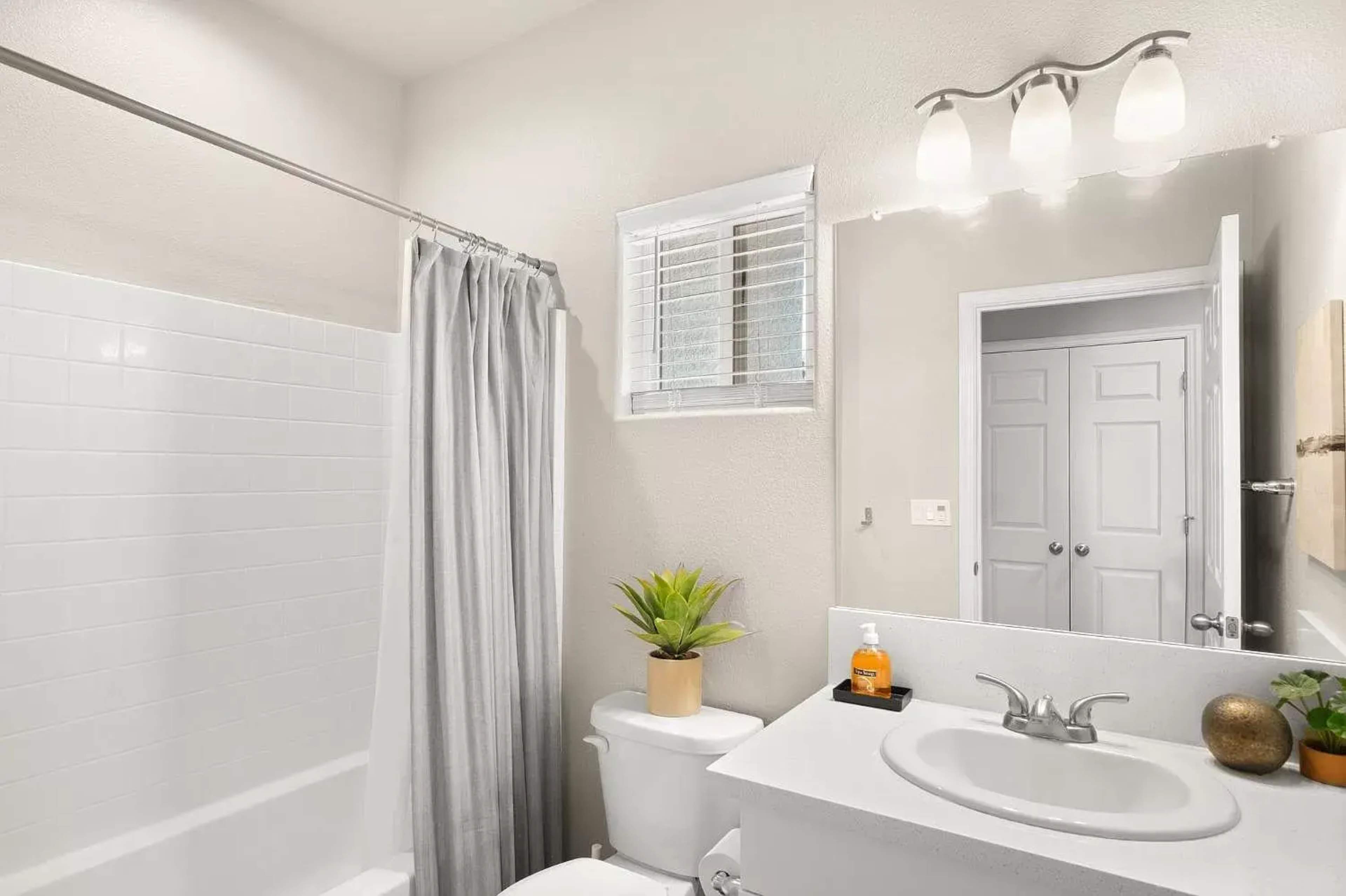 A clean bathroom features a bathtub with a shower curtain, a sink with a faucet, a mirror, and a plant on the counter.