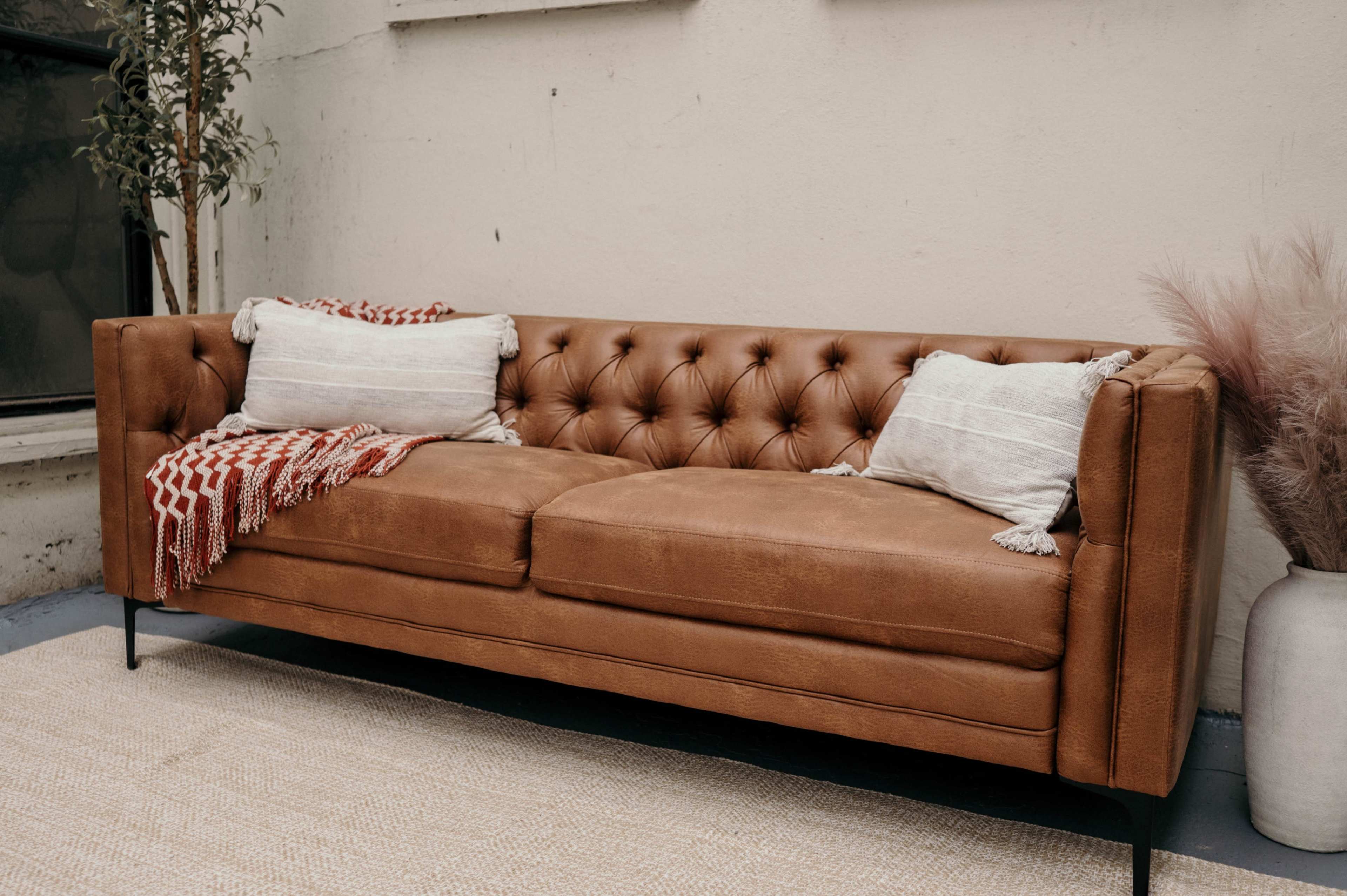 A brown leather tufted sofa with two decorative pillows is placed against a neutral wall next to a decorative plant and a light-colored rug.