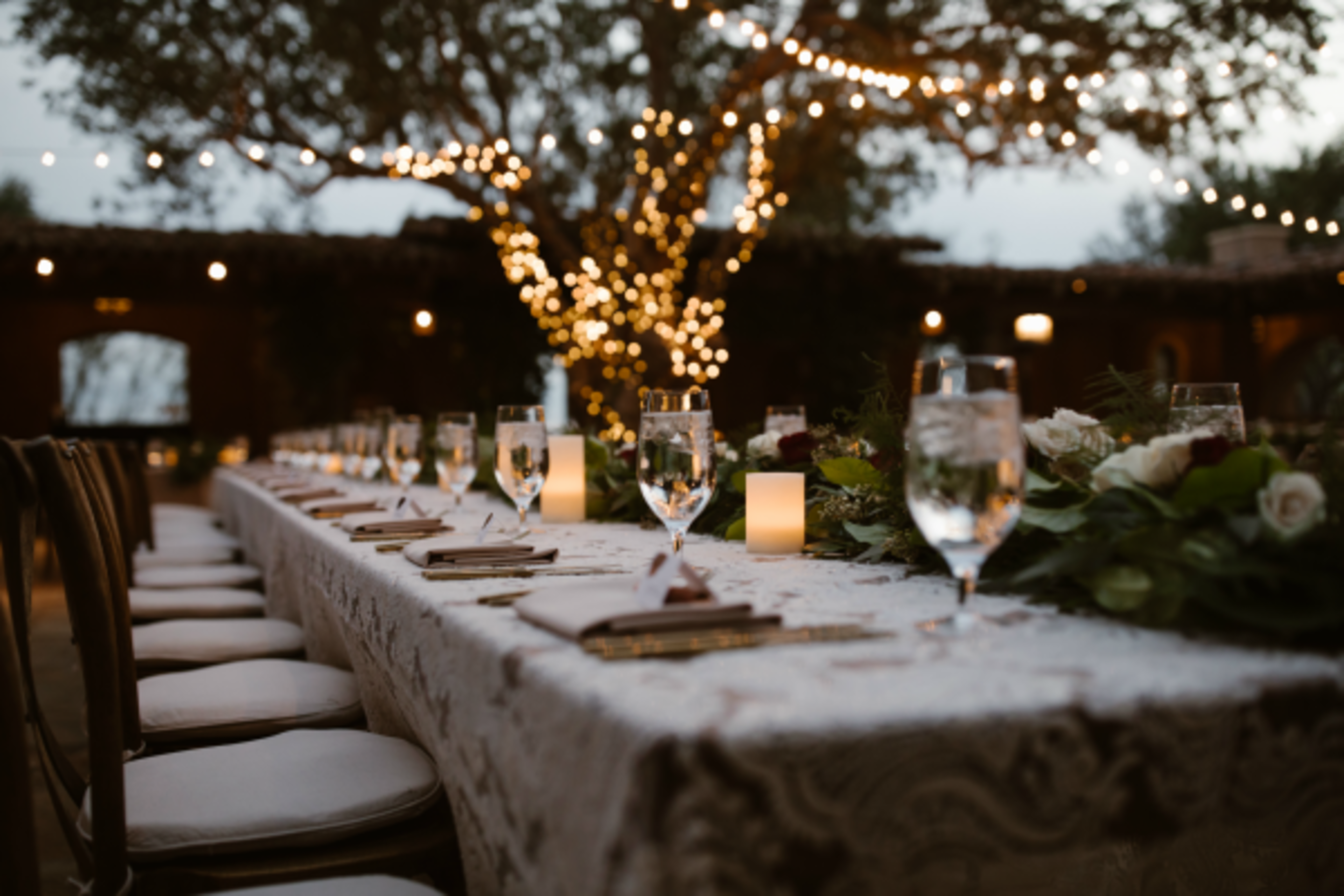 A long dining table is elegantly set outdoors, adorned with glasses of water, candles, and a floral centerpiece, with fairy lights strung overhead.