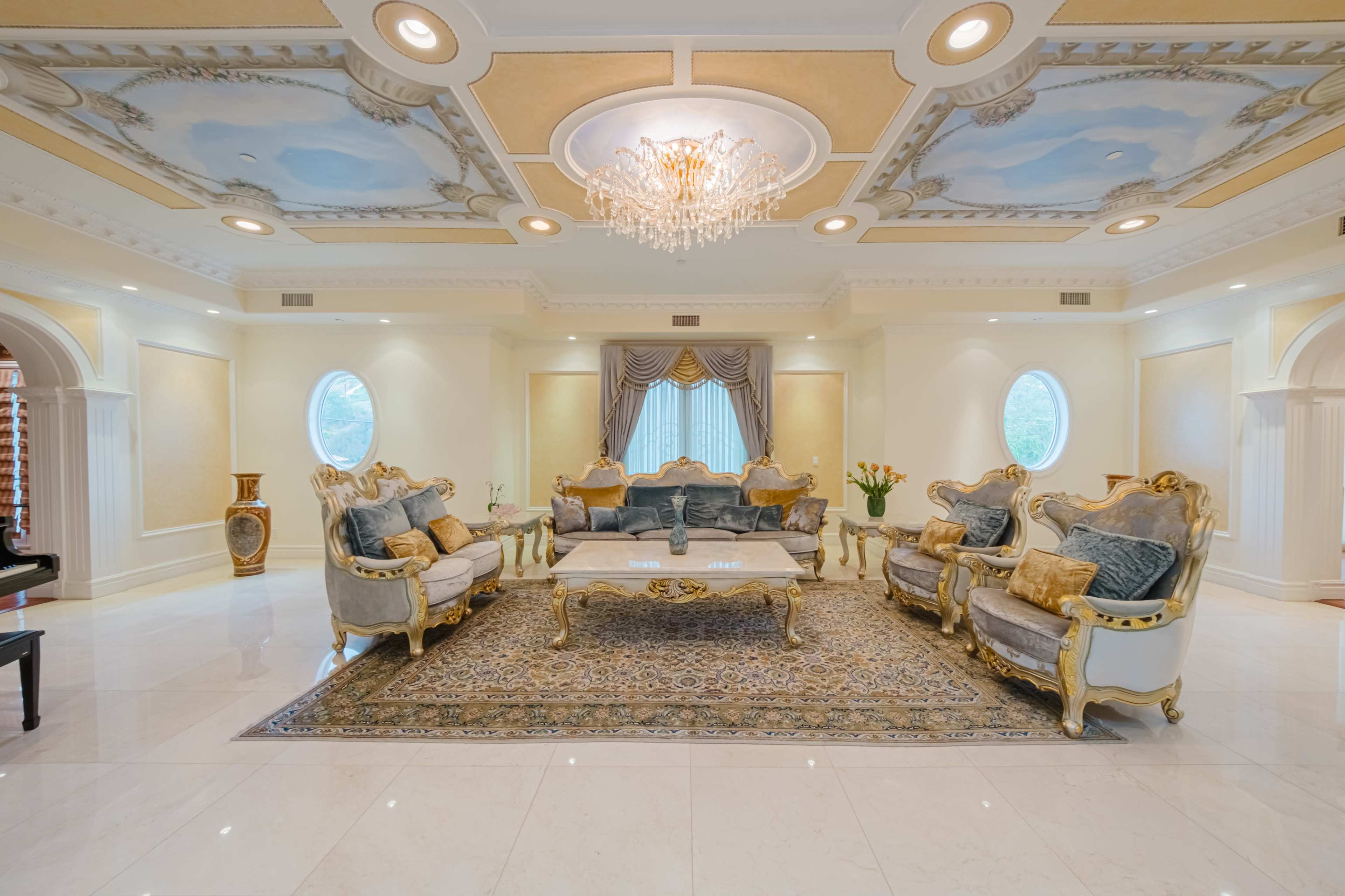 A luxurious living room with ornate gold-trimmed furniture, a chandelier, and a detailed ceiling, featuring round windows and a large area rug.
