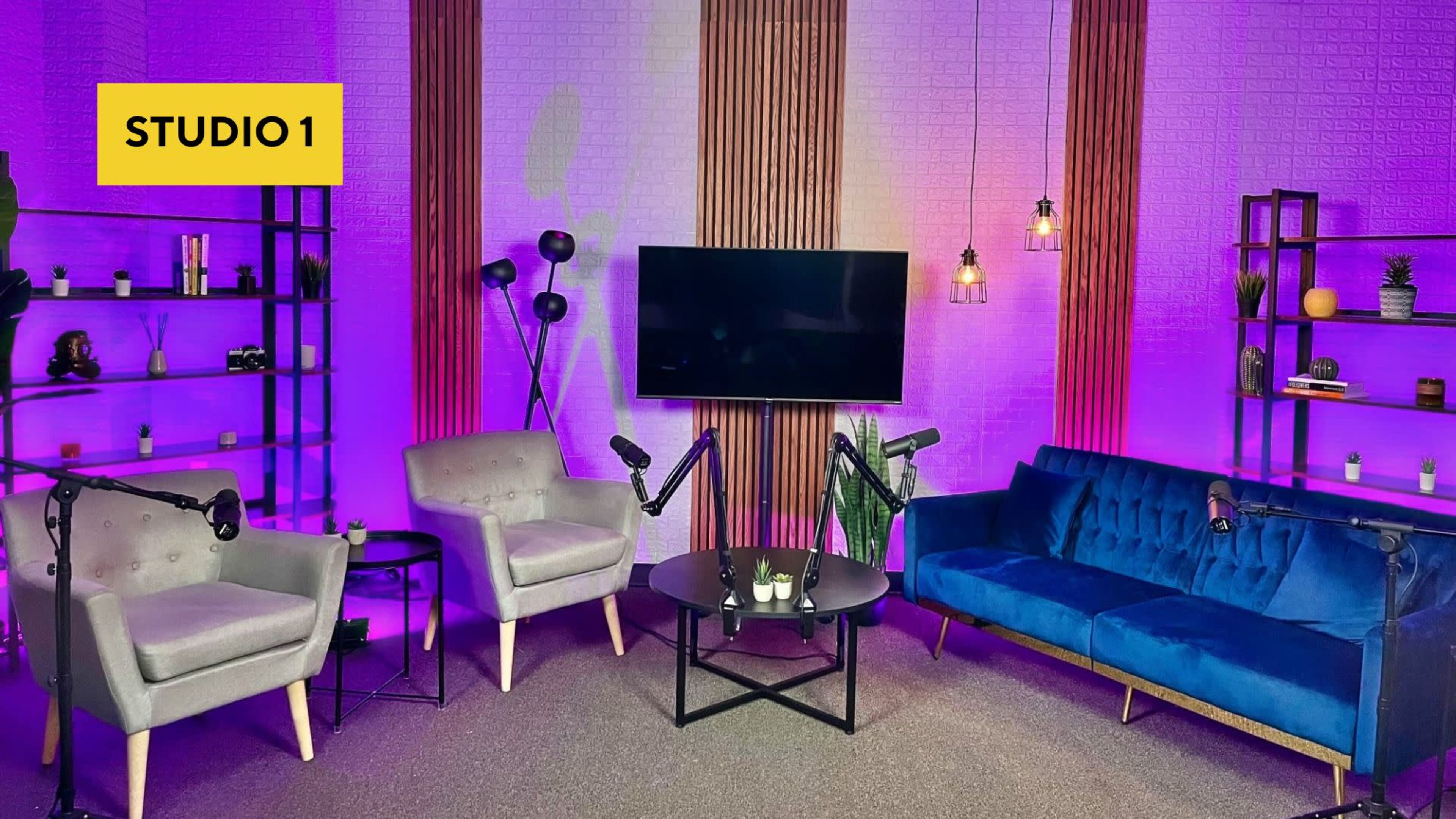 A modern studio setup with purple lighting, featuring a blue velvet couch, two armchairs, a coffee table, and a wall-mounted television.