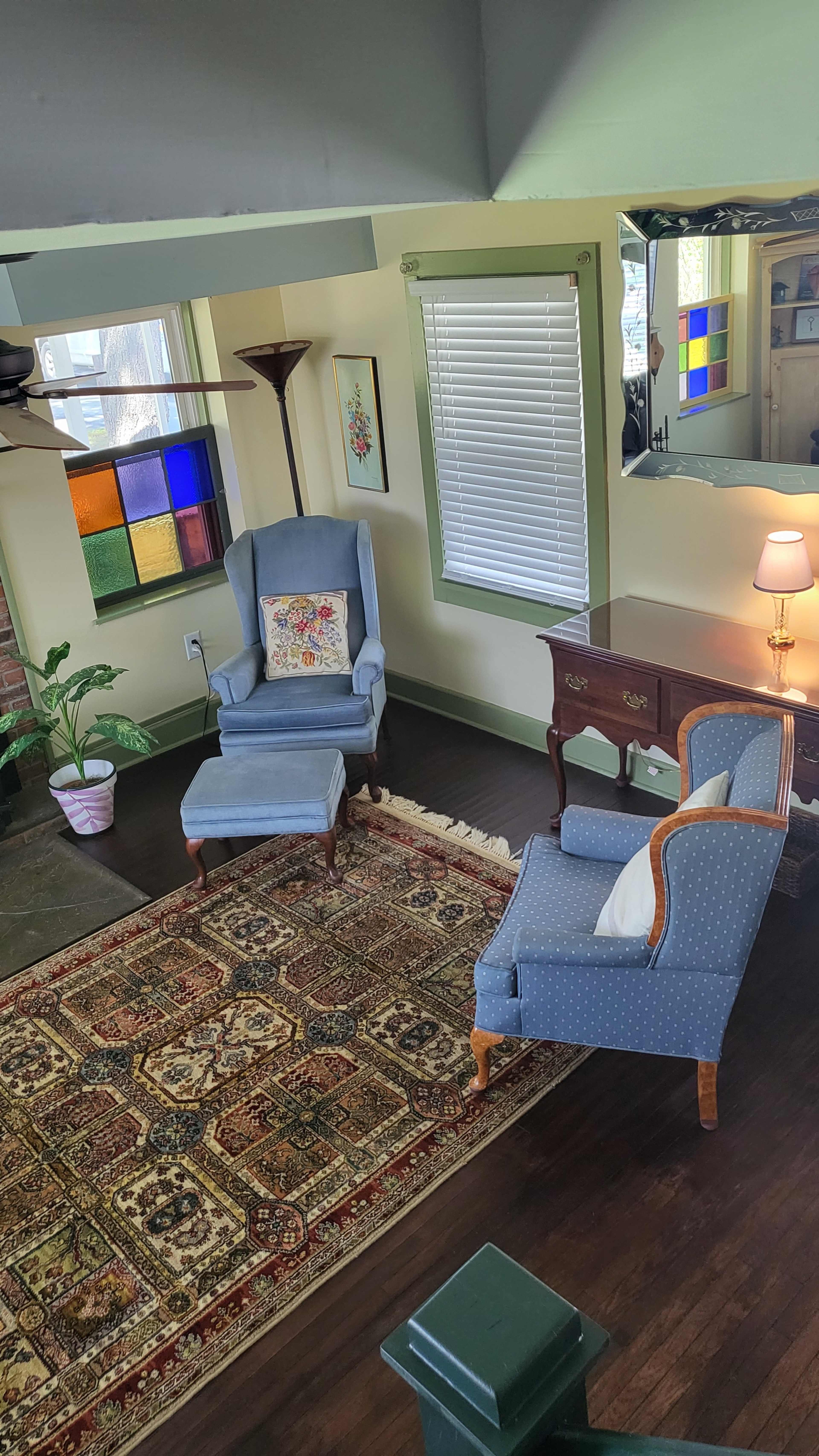 A cozy living room with two upholstered chairs, a wooden side table, and a decorative rug, set against light green walls and a stained glass window.