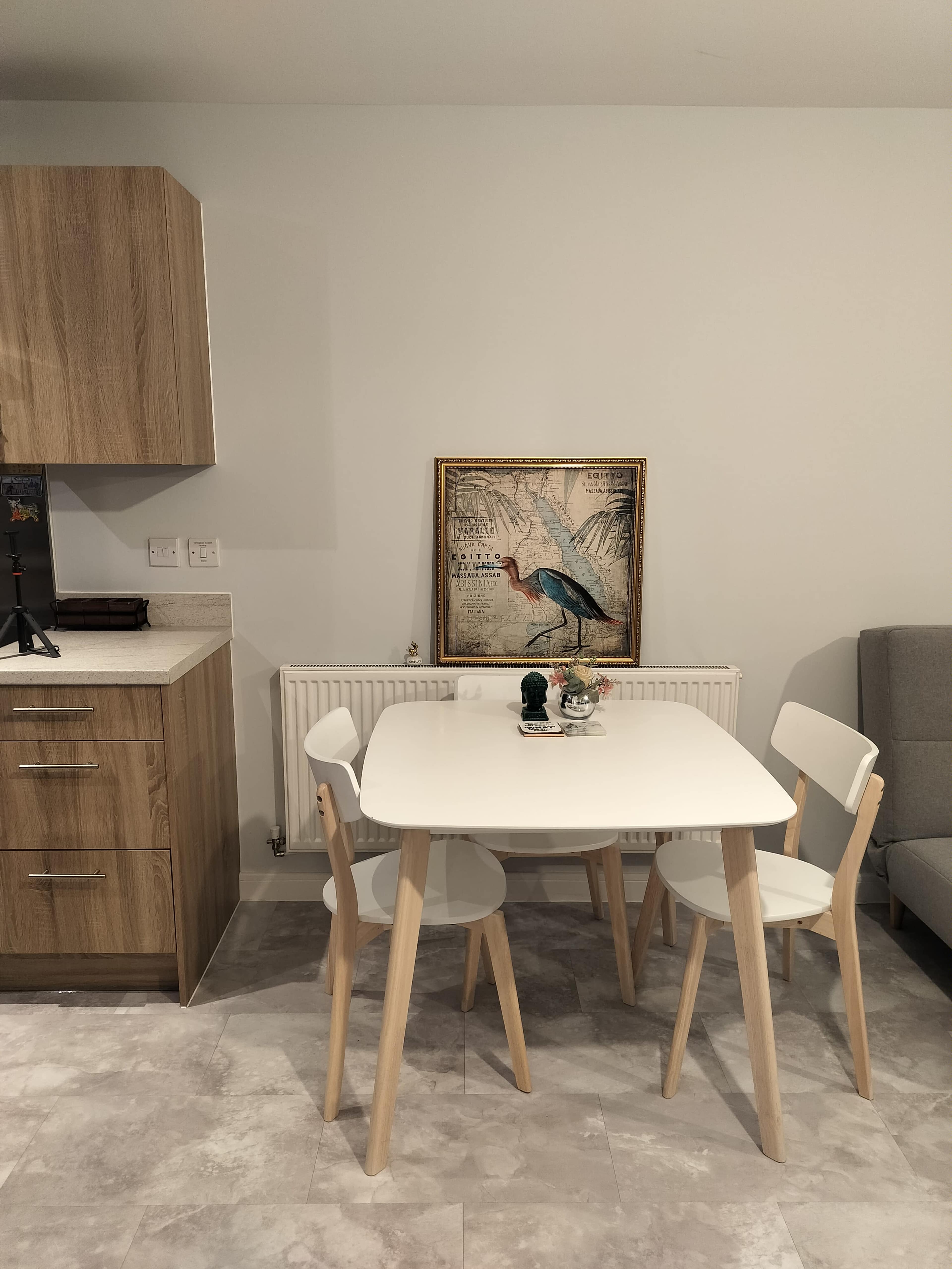 The image shows a minimalist dining area with a white table and three chairs, adjacent to a wooden kitchen cabinet and a framed bird painting on the wall.
