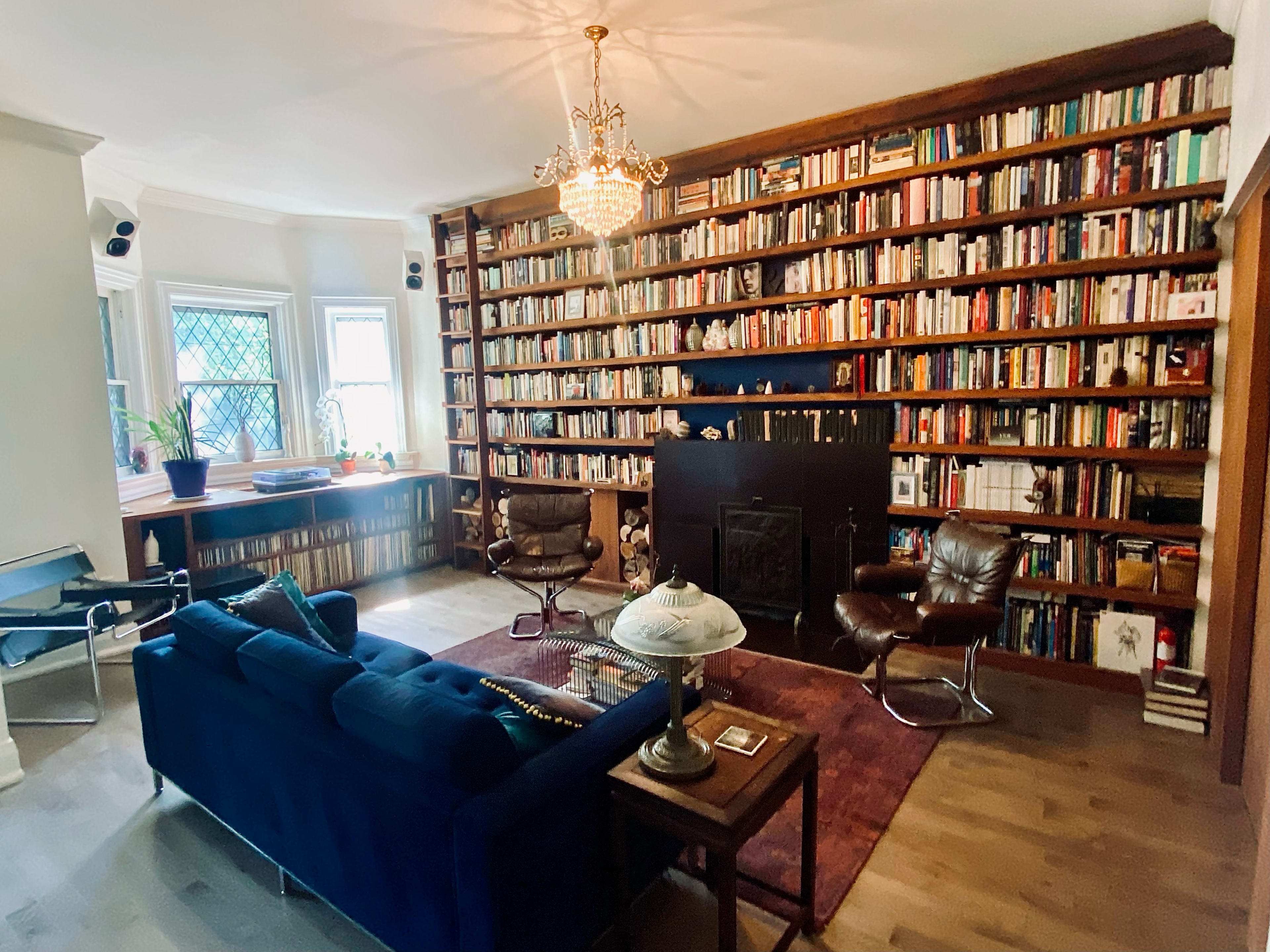 A cozy reading room with a large bookshelf filled with books, a dark sofa, two leather chairs, and a lamp on a wooden side table.